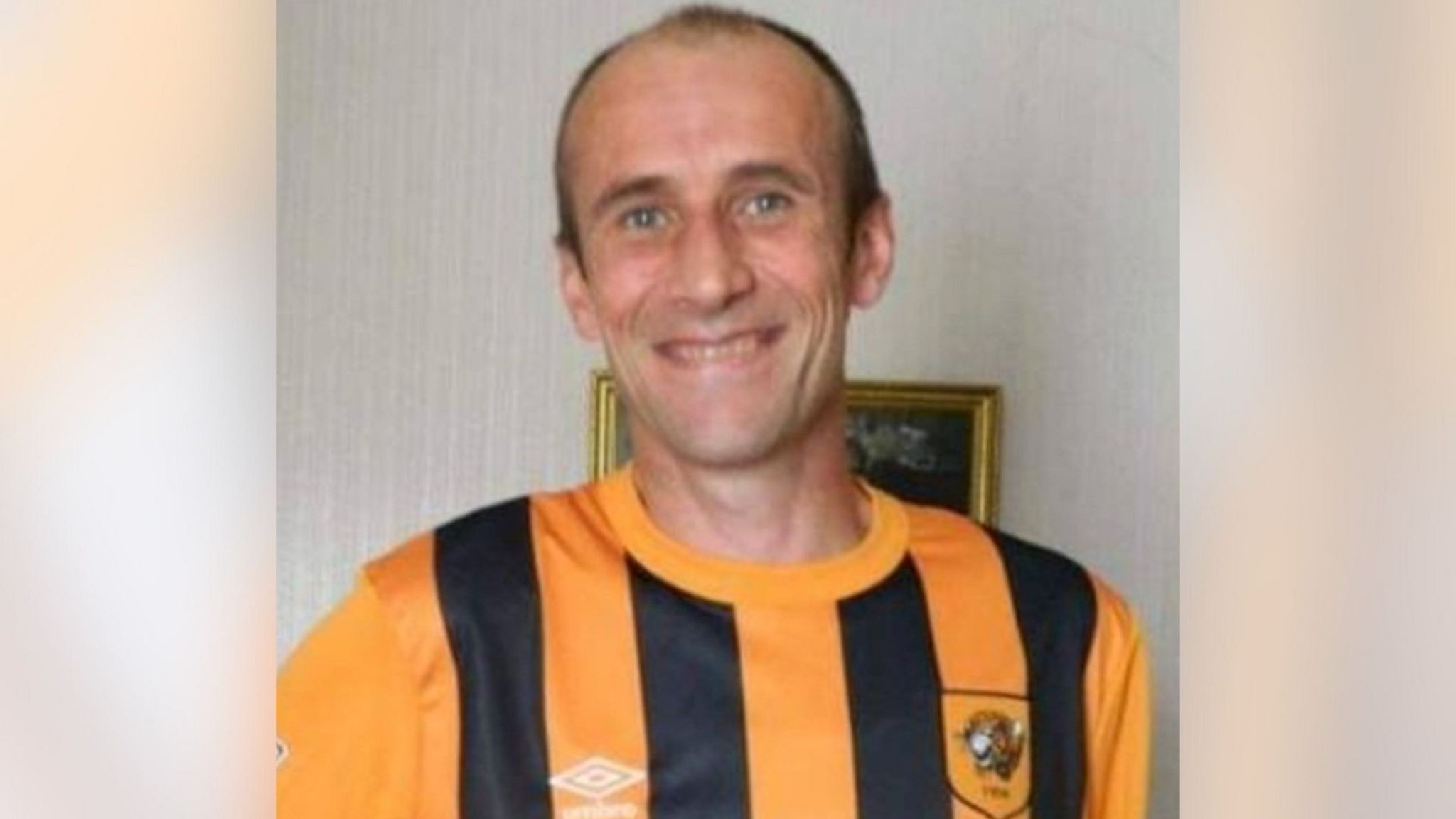 Christopher King, smiling and wearing a yellow and black striped Hull City football shirt. He is standing in front of a painting. 