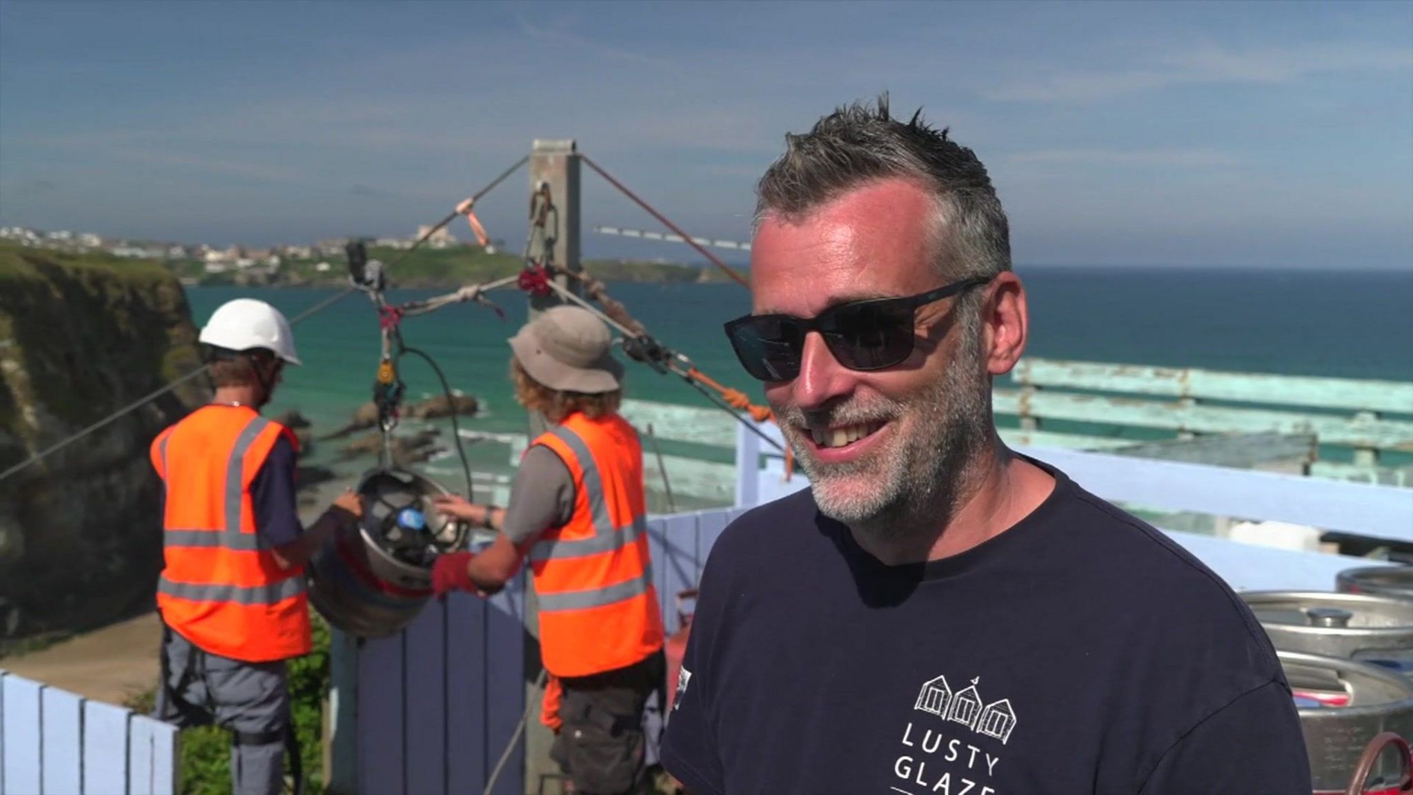 A photo of Robin Parris. It shows two people behind him in hi-vis jackets pushing the beer keg down the zipline. Mr Parris is in the forefront wearing sunglasses and a T-shirt saying Lusty Glaze