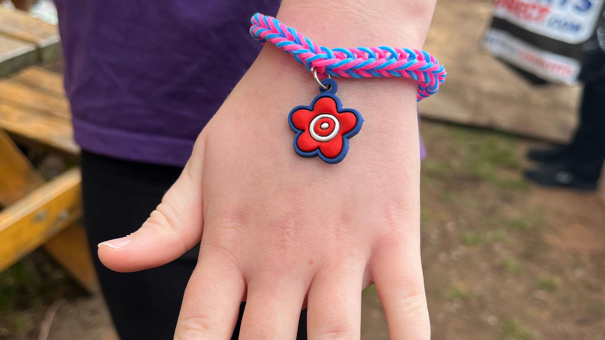 A child's wrist with a bracelet around it. The bracelet is pink and blue, made out of elastic, with a red, white and blue flower charm attached to it