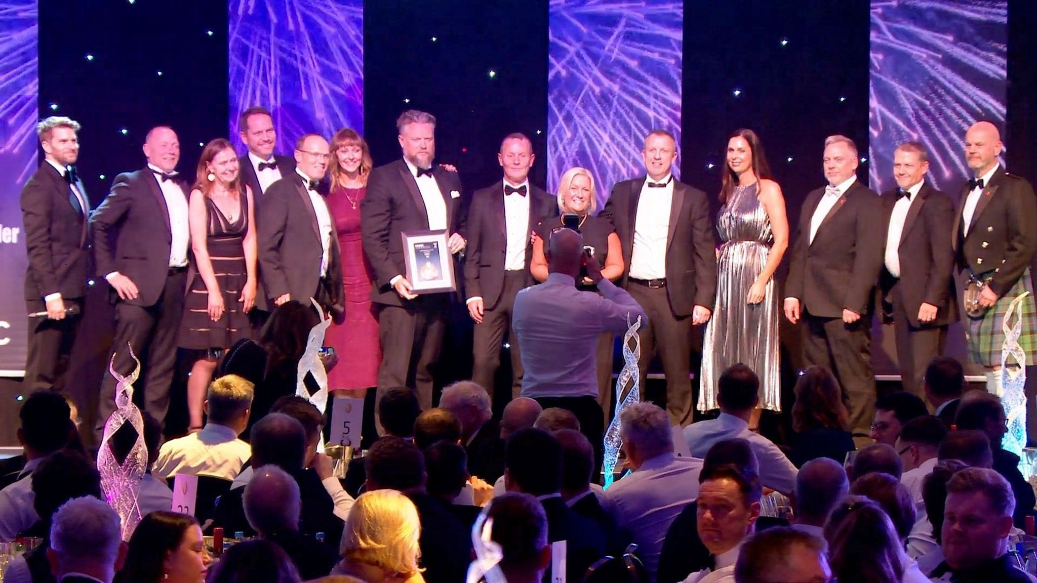 More than a dozen people on stage posing for a picture as they accept an award. The dress code is black tie. A phographer in a white shirt is standing in front of the stage with his camera raised and other guests are seated at tables in the foreground.