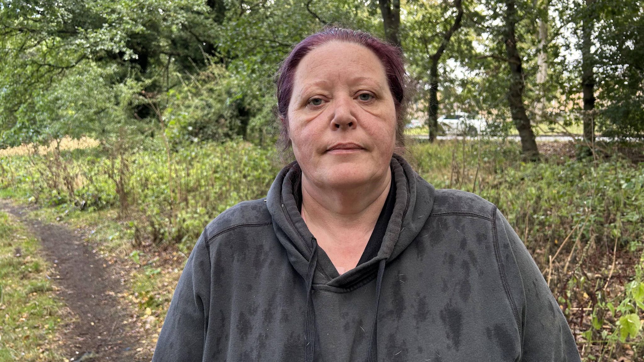 A woman with dyed purple hair wearing a black hoodie is stood on a rural path where she walks her dogs. She is surrounded by trees and it is raining, with rain drops on her hoodie.