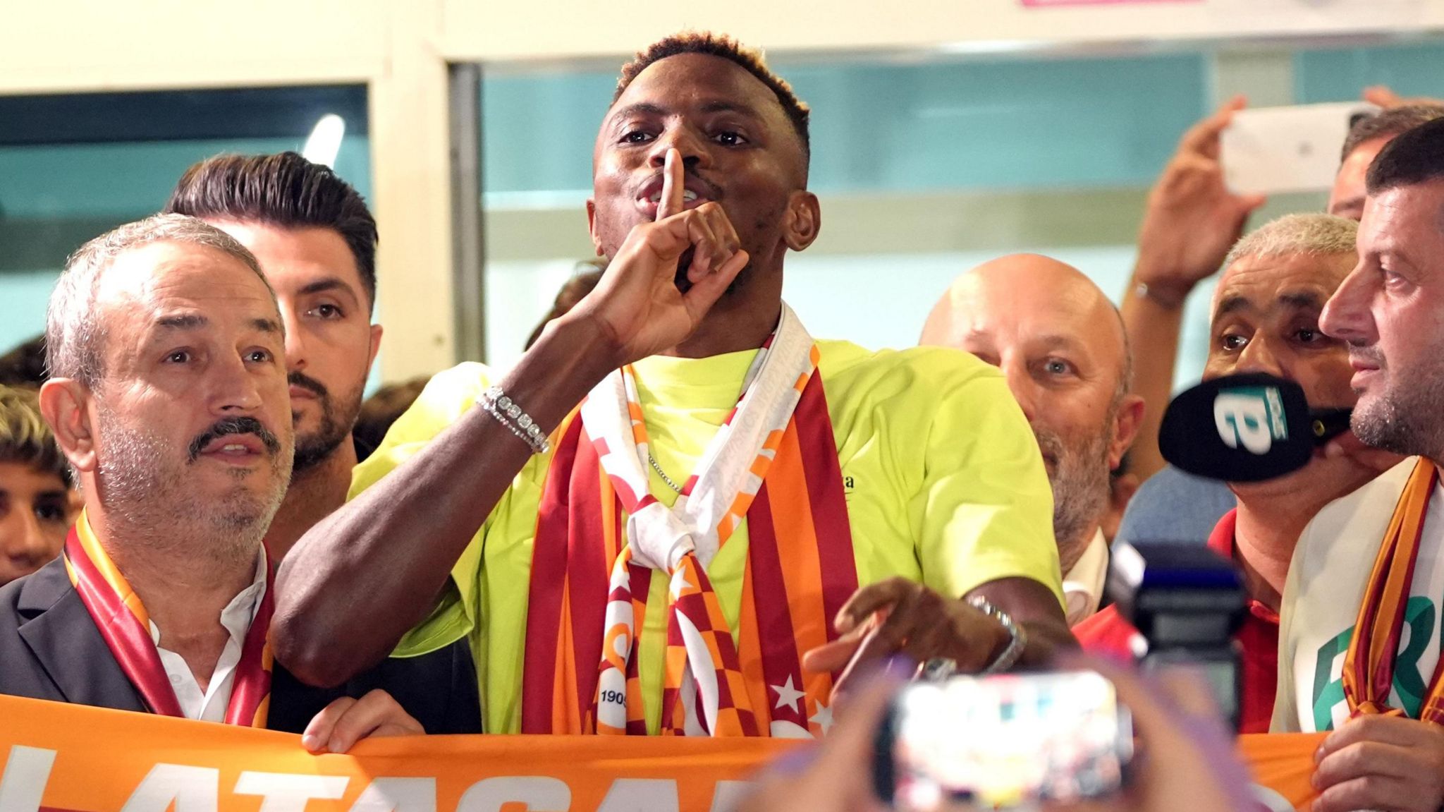 Victor Osimhen wearing Galatasaray colours around his neck holds his finger up to his lips in a shush gesture as he is surrounded by fans on his arrival in Instanbul