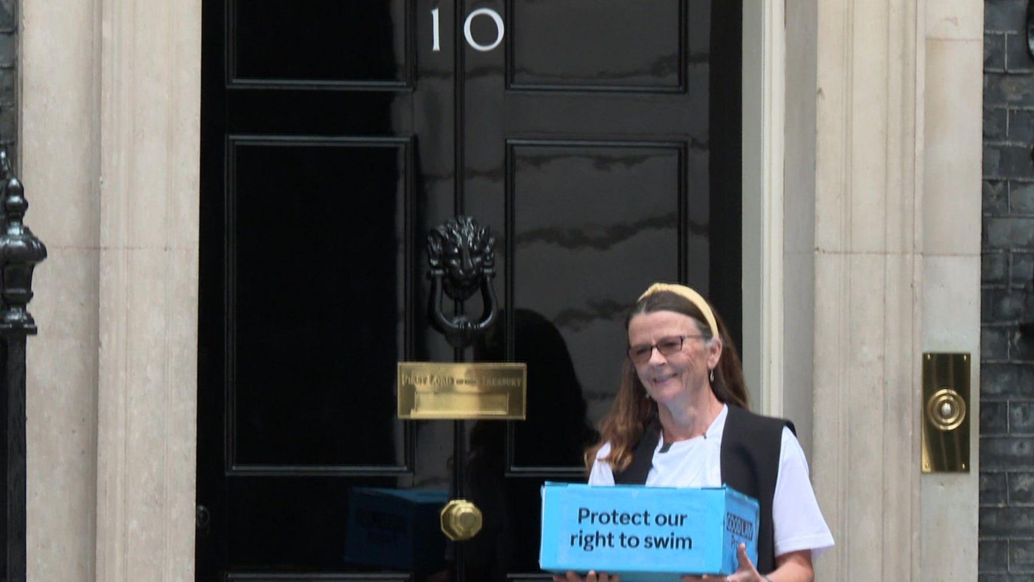 Jo Bateman with her petition contained inside a blue box with the words "protect our right to swim" written on it in black letters. She is standing in front of 10 Downing Street's front door.