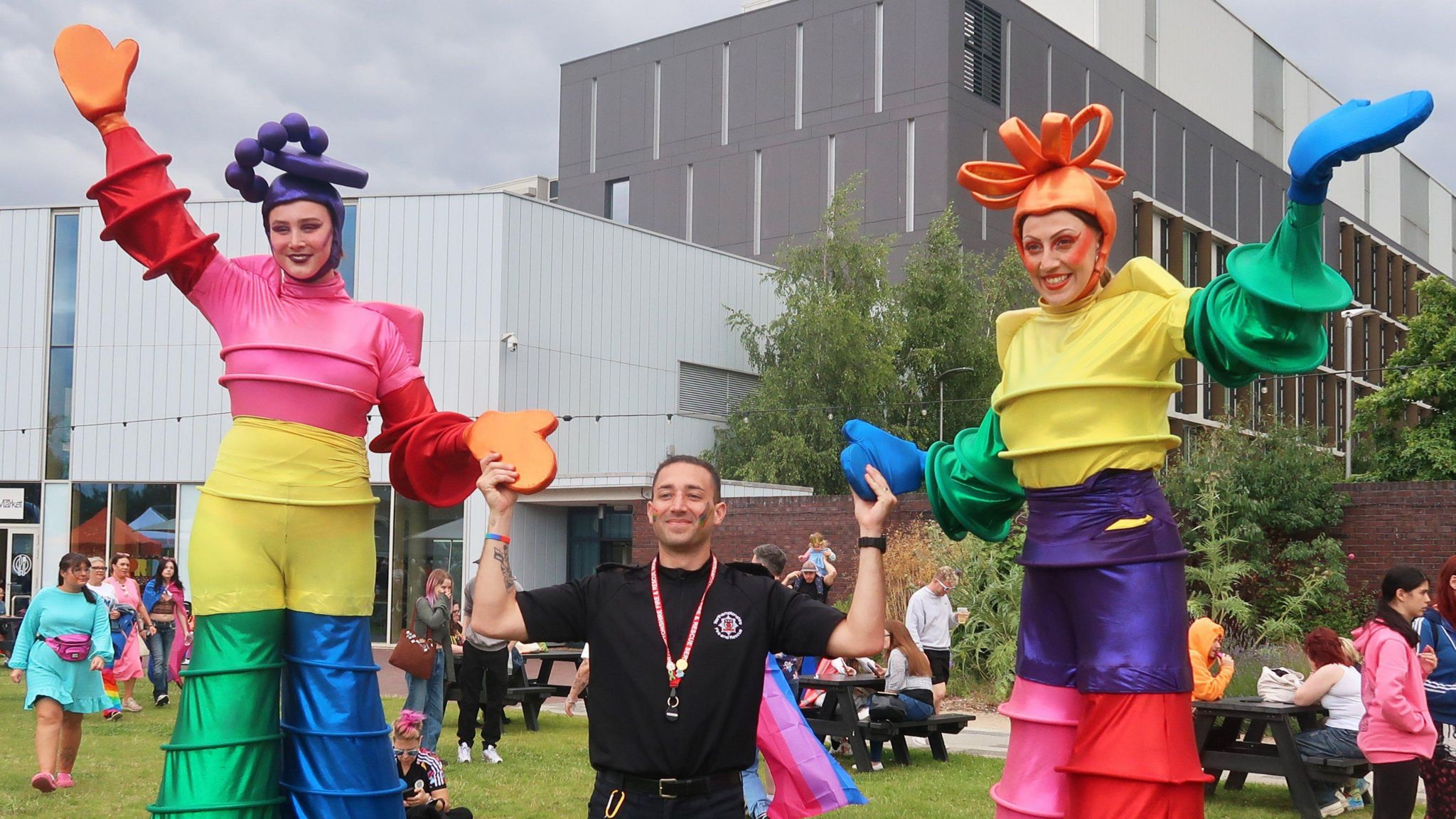 Two stilt walkers in colourful costumes hold hands with a man not on stilts