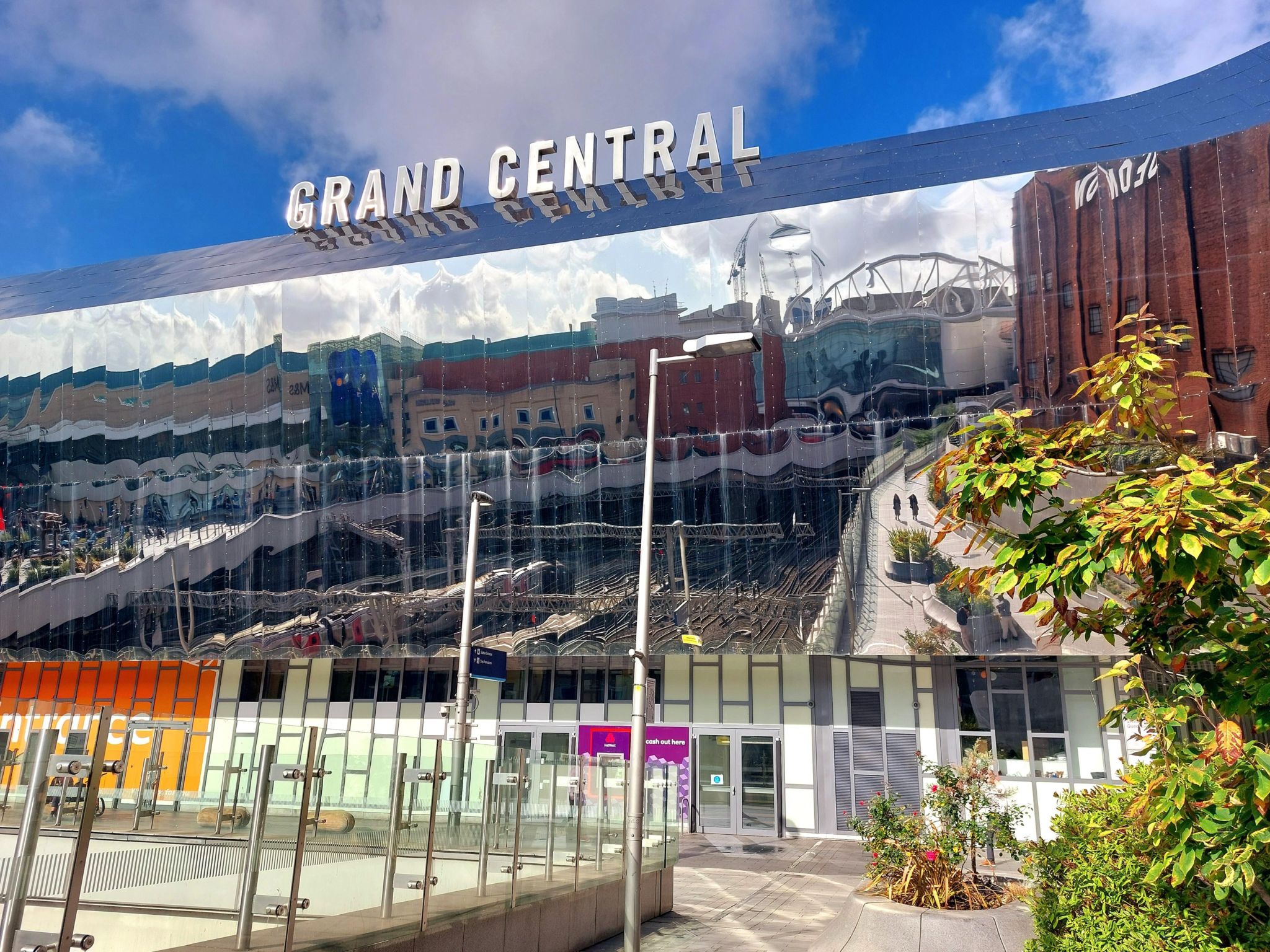 Reflective panels on the front of the shopping centre show a reflection of the city skyline opposite. The sky is blue with light white clouds. 