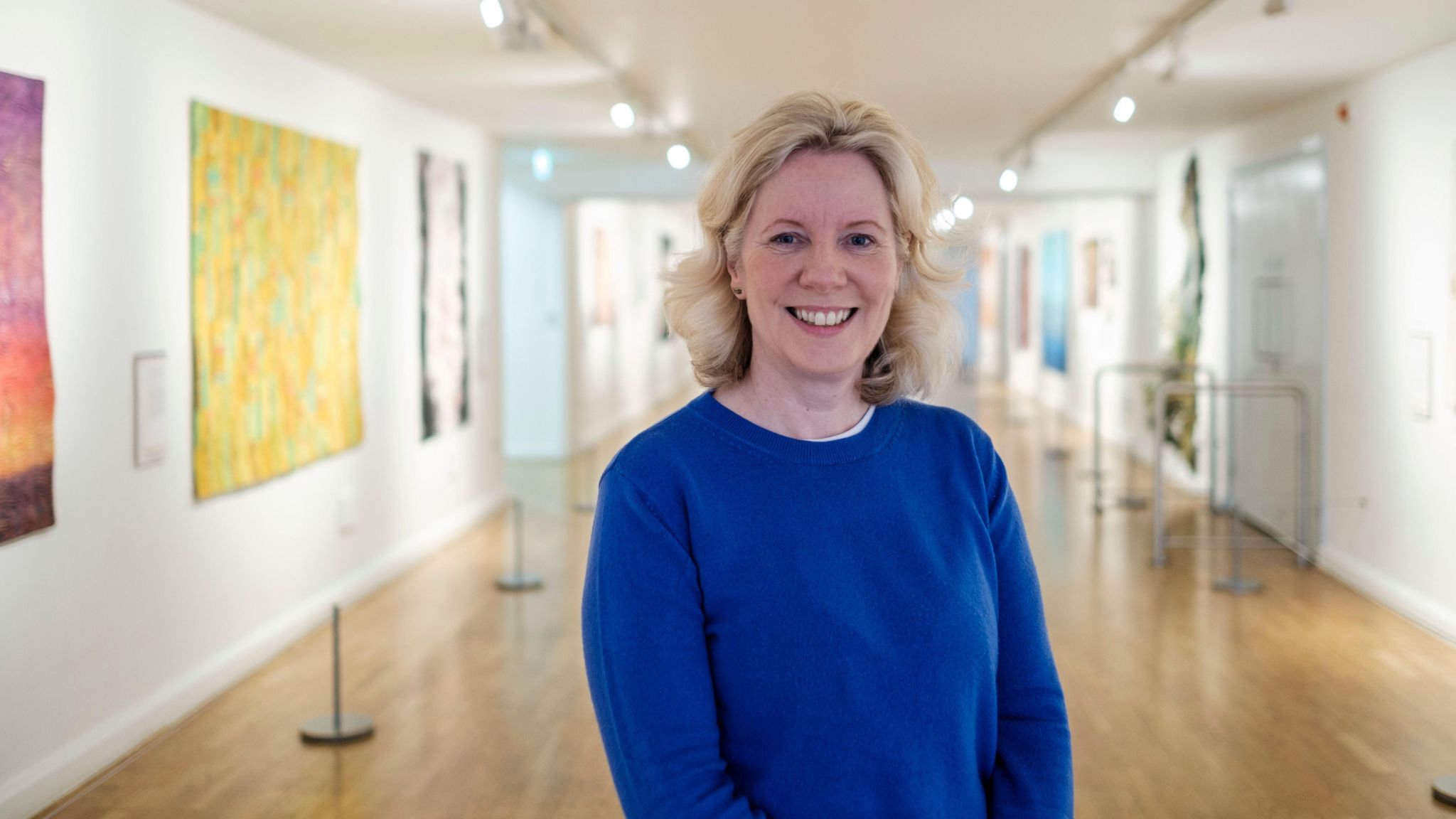 Debbie Kermode wearing a blue top and standing among paintings in a gallery