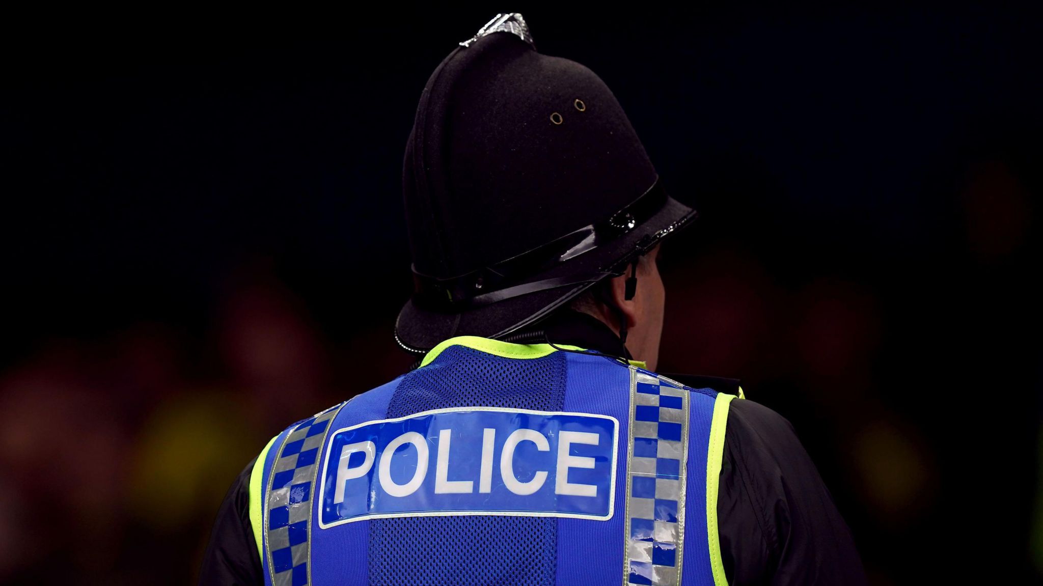 A police office with his back to the camera wearing a helmet and high-vis jacket with 'police' written on it