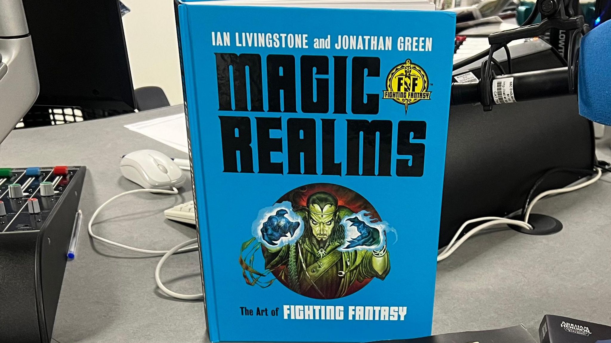 The Magic Realms The Art of Fighting Fantasy book stood up on a desk. It is a blue book with big, black letters. 