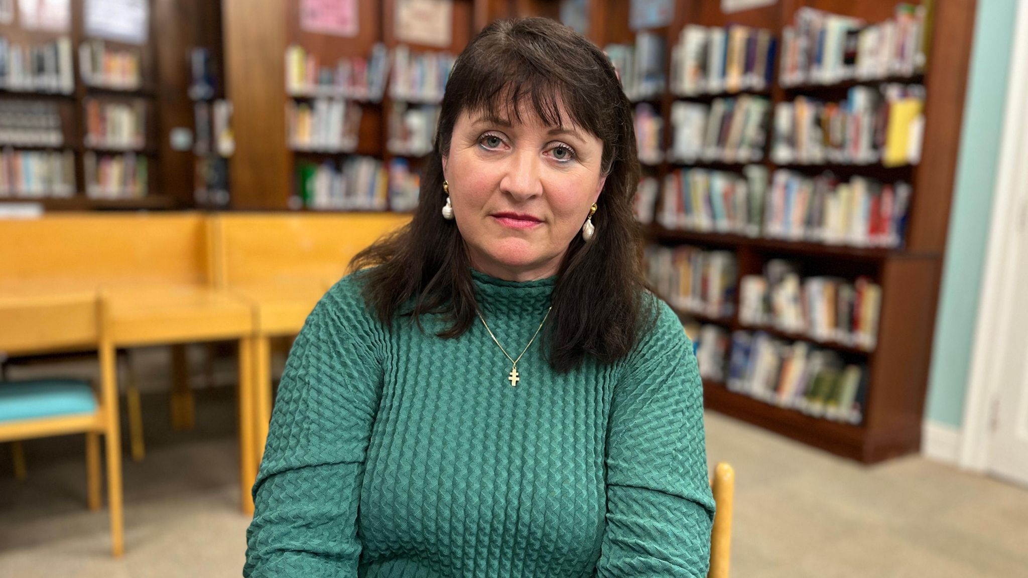 Katherine Jeffrey sits in a school library wearing a green dress, pearl earrings and gold cross necklace