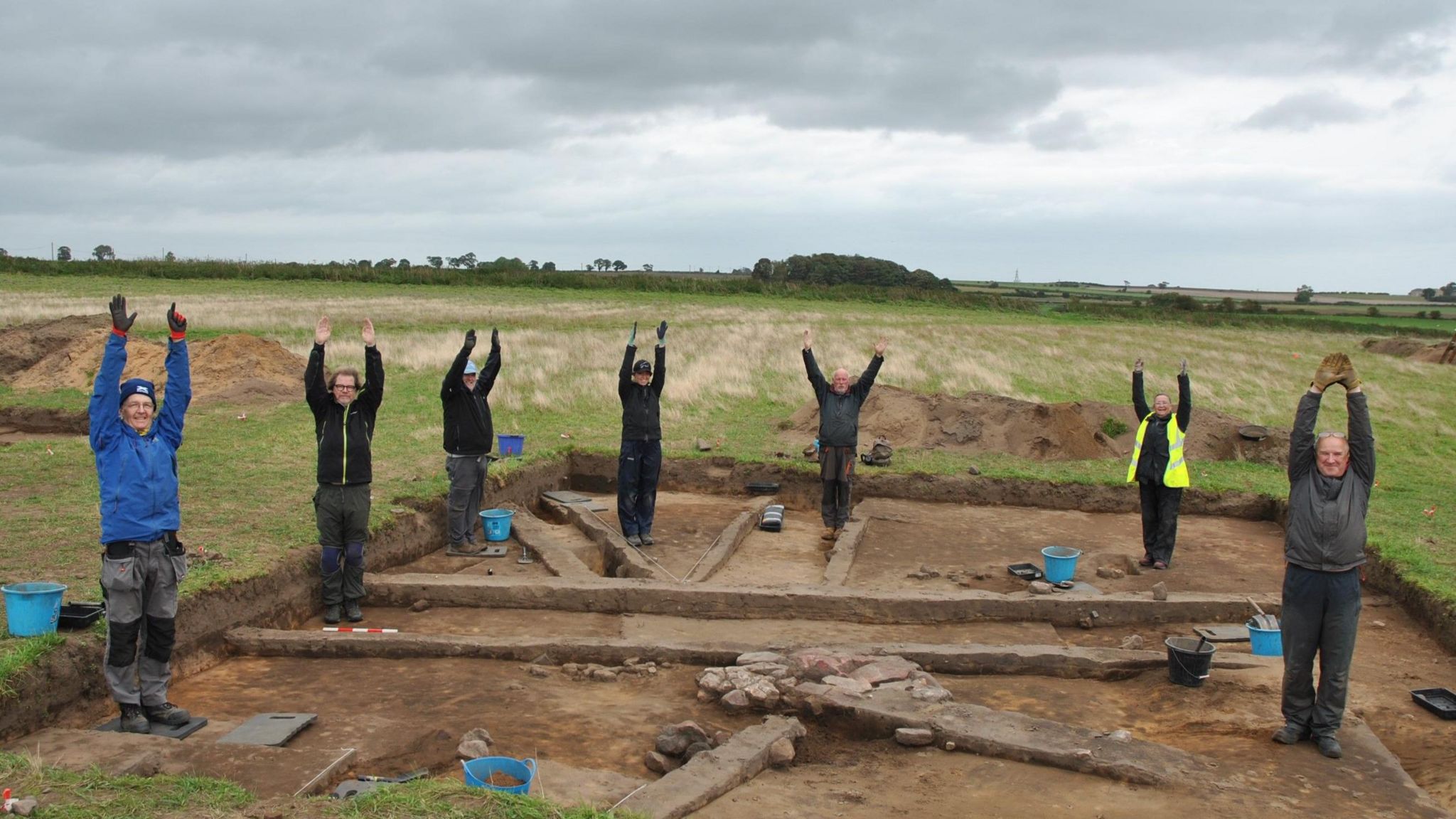A number of volunteer archaeologists standing in the site of a building holding their arms in the air