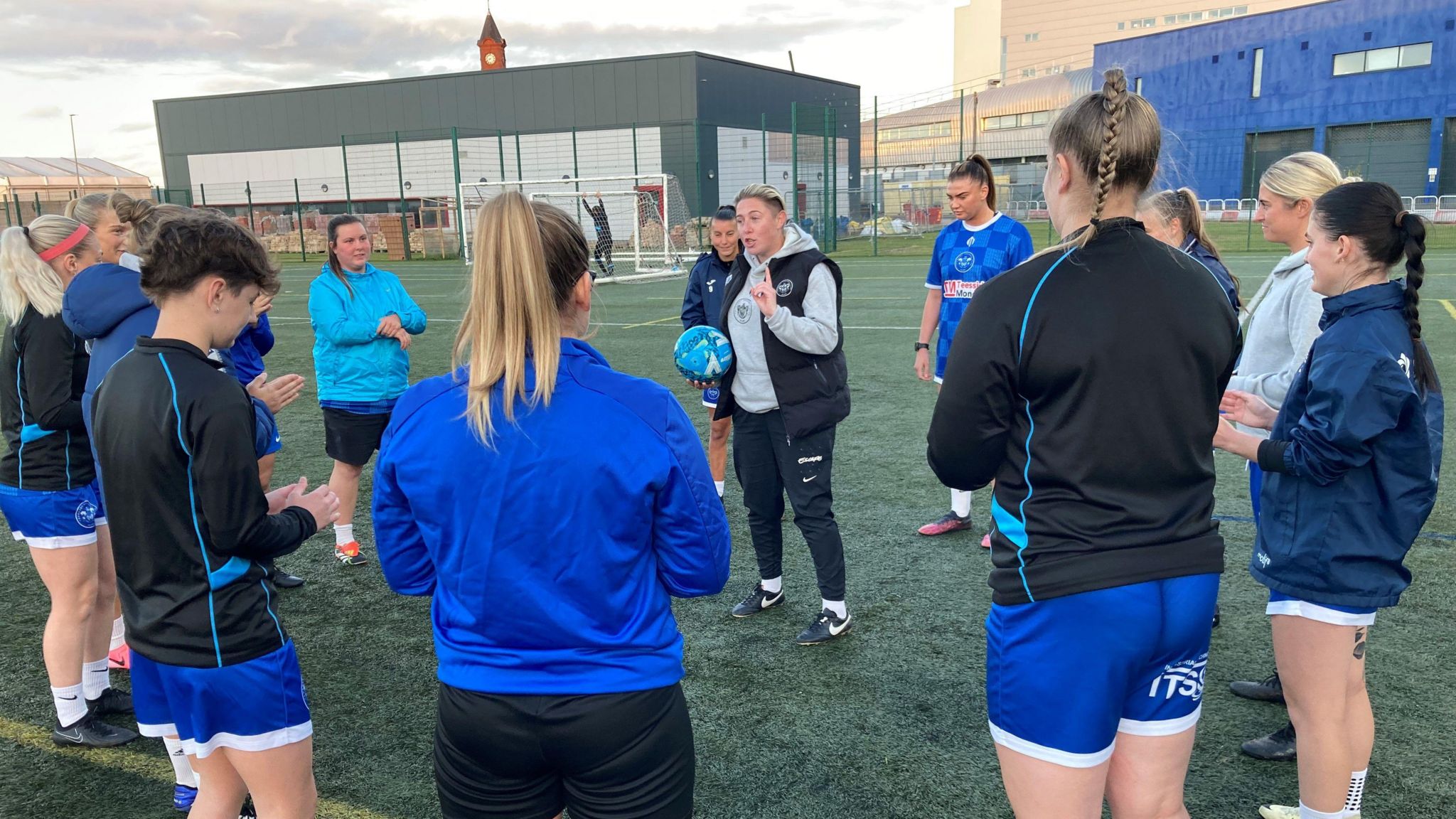 Co-Manager Abbey Lyle holding a ball and talking to Thornaby FC players who have formed a circle around her on the football pitch. 
