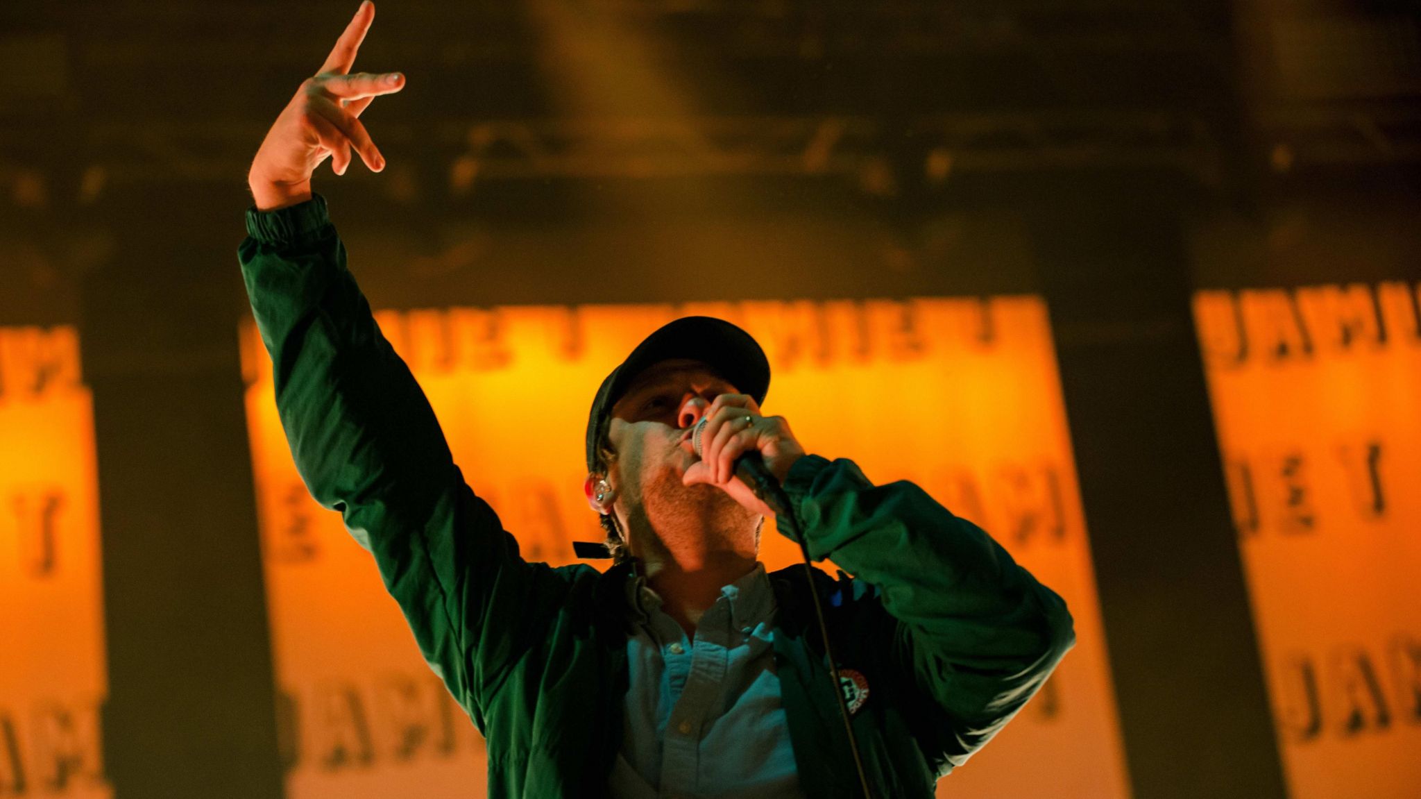 Jamie T sings into a microphone, a hand in the air, wearing a jacket and cap.