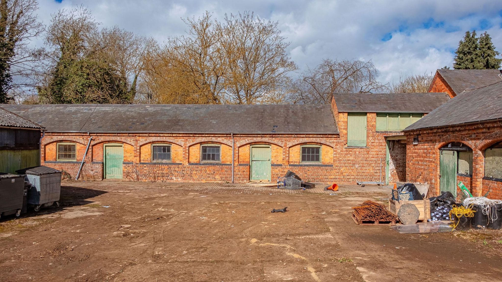 A dusty yard with a red-brick stable building with green doors in the backdrop.