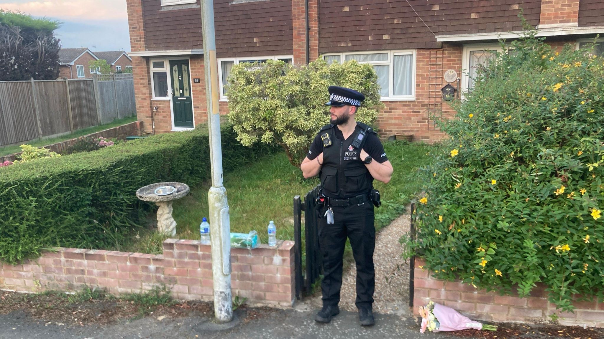 A police officer stands outside a house in Bright Ridge and a floral tribute has been left on the pavement