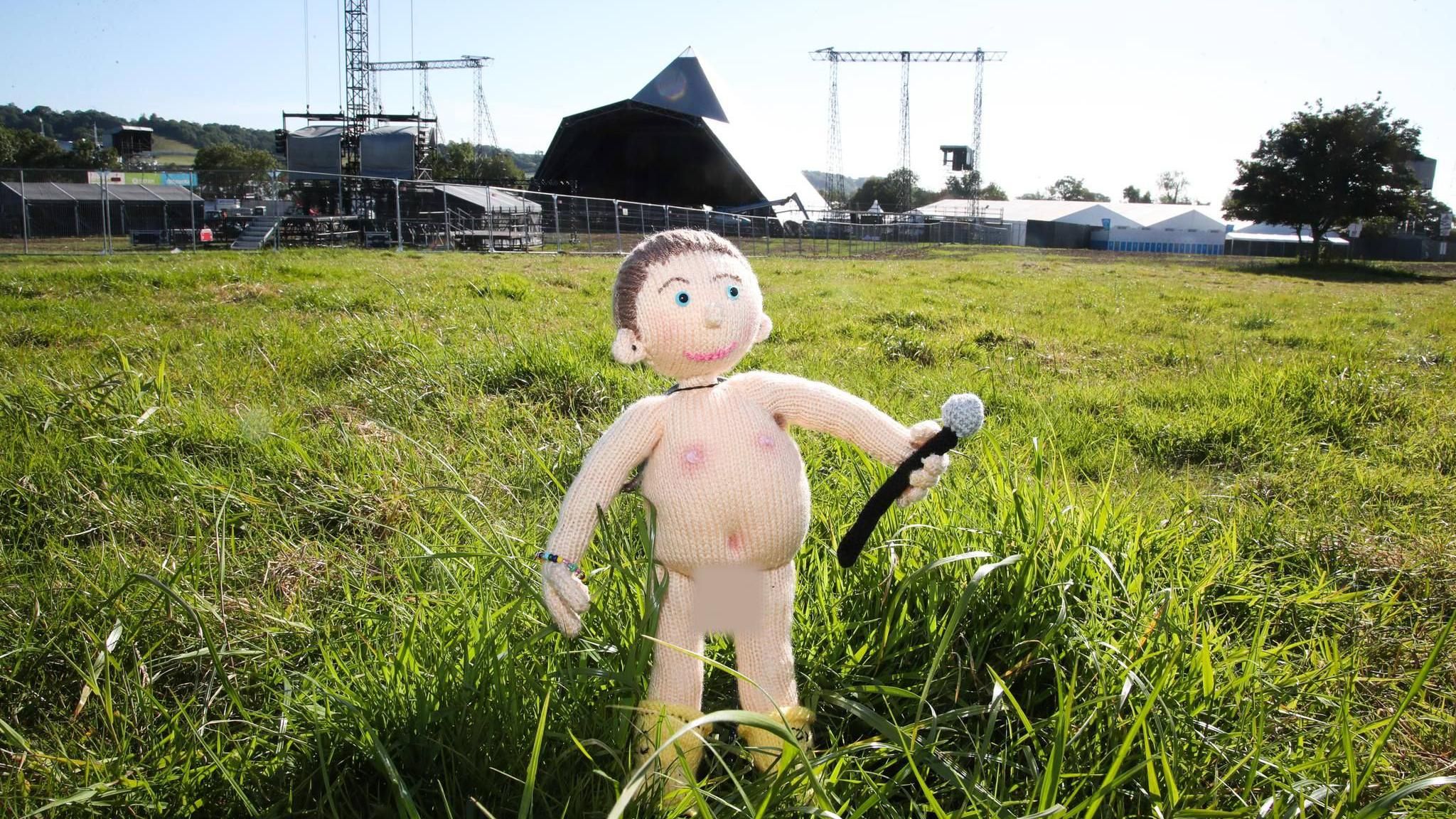 A knitted doll resembling Chris Martin standing on the grass in front of Pyramid Stage at Glastonbury. The doll is naked and holding a microphone