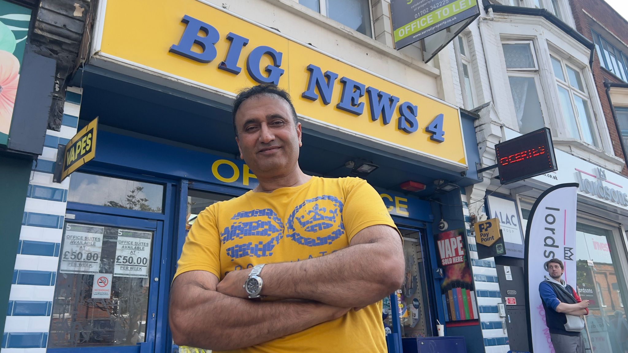 Mohammad Imran smiling and looking down at the camera with his arms crossed. He is standing in front of his shop which has a sign reading "Big News 4"