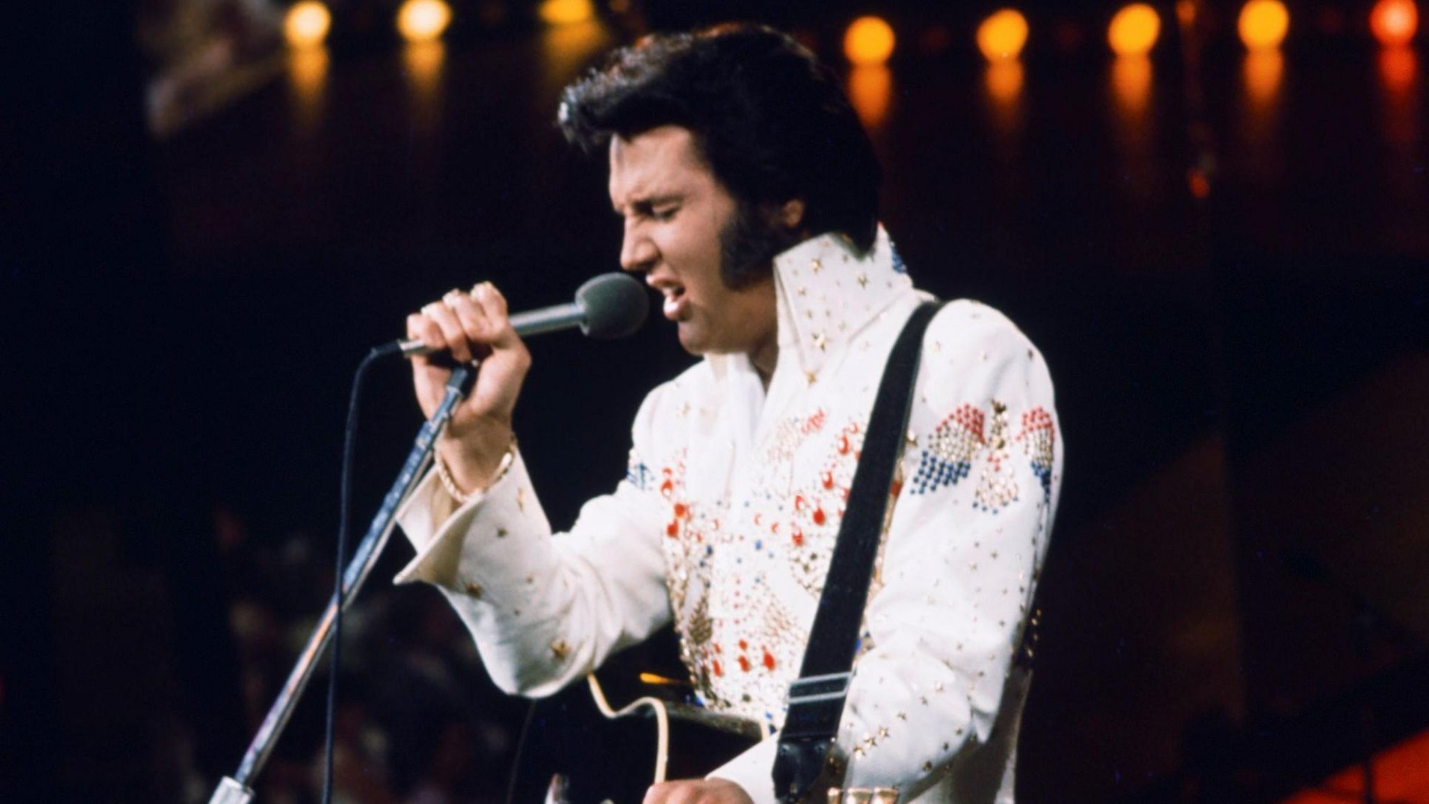 Elvis Presley sings on stage. He is wearing a white sparkly suit and is playing a black acoustic guitar
