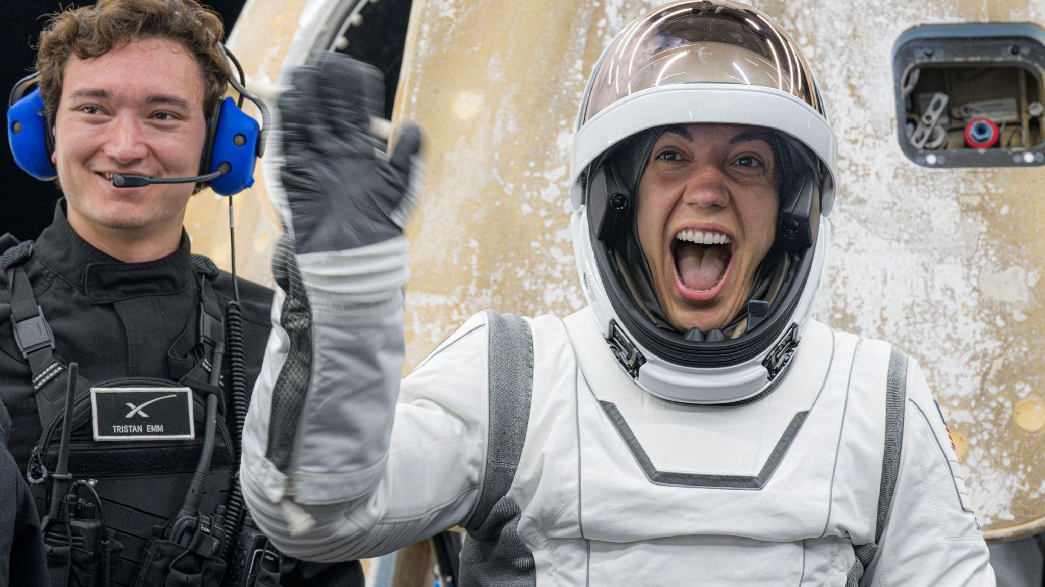 An astronaut waves at the crowd. She's smiling broadly and wearing her white-and-grey spacesuit and is standing infront of the Dragon capsule.