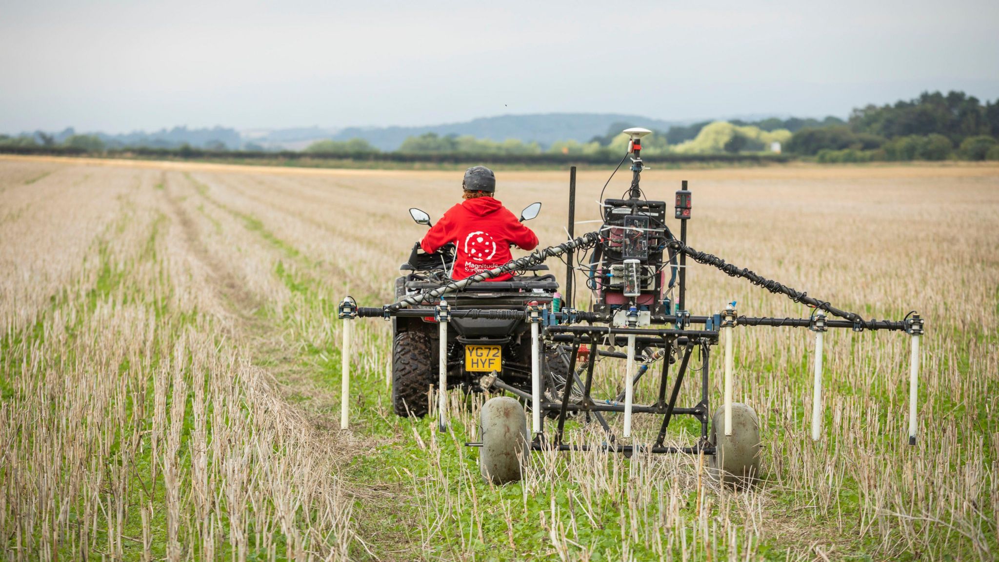 A person in a red top on a quad bike towing survey equipment across a field. The equipment is made up of several metal poles with some dangling over the field and mounted on two wheels.