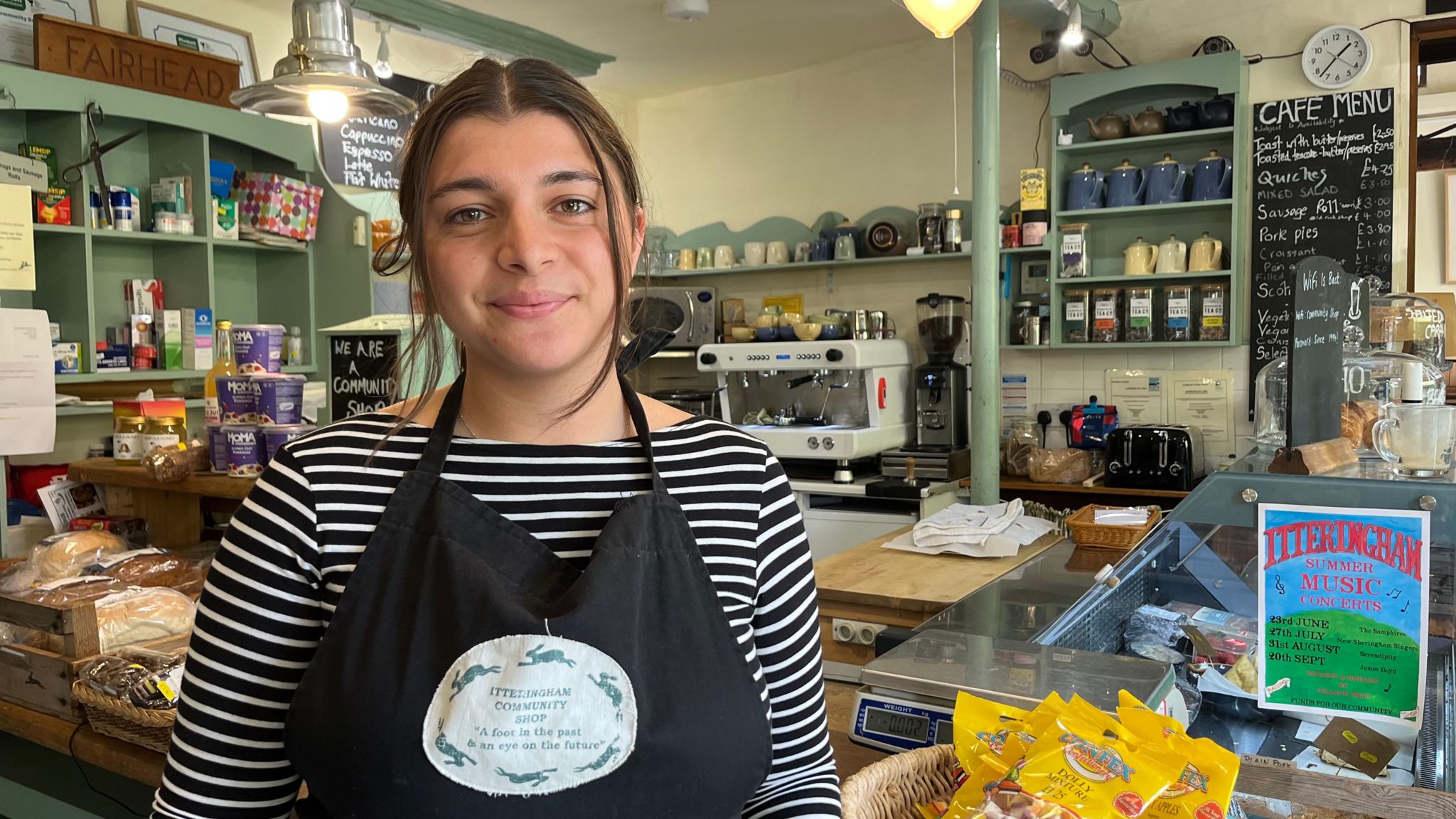 A smiling Rosie Hull looks directly at the camera, standing in her community shop and wearing a black and white striped top and black apron. 