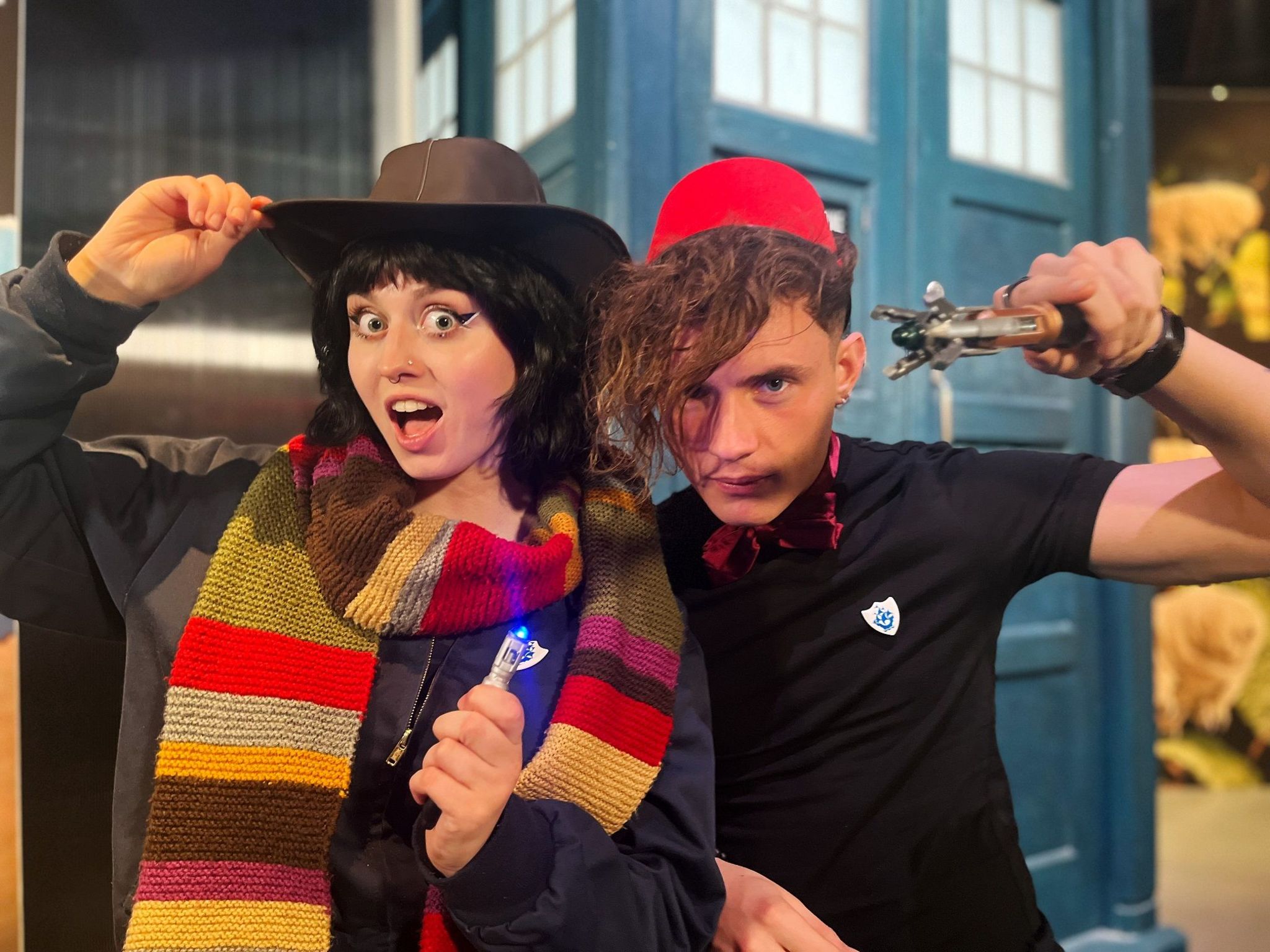 Abby Cook, wearing a stripy scarf and large hat, with Joel Mawhinney. Both are holding sonic screwdrivers. There is a Tardis in the background.