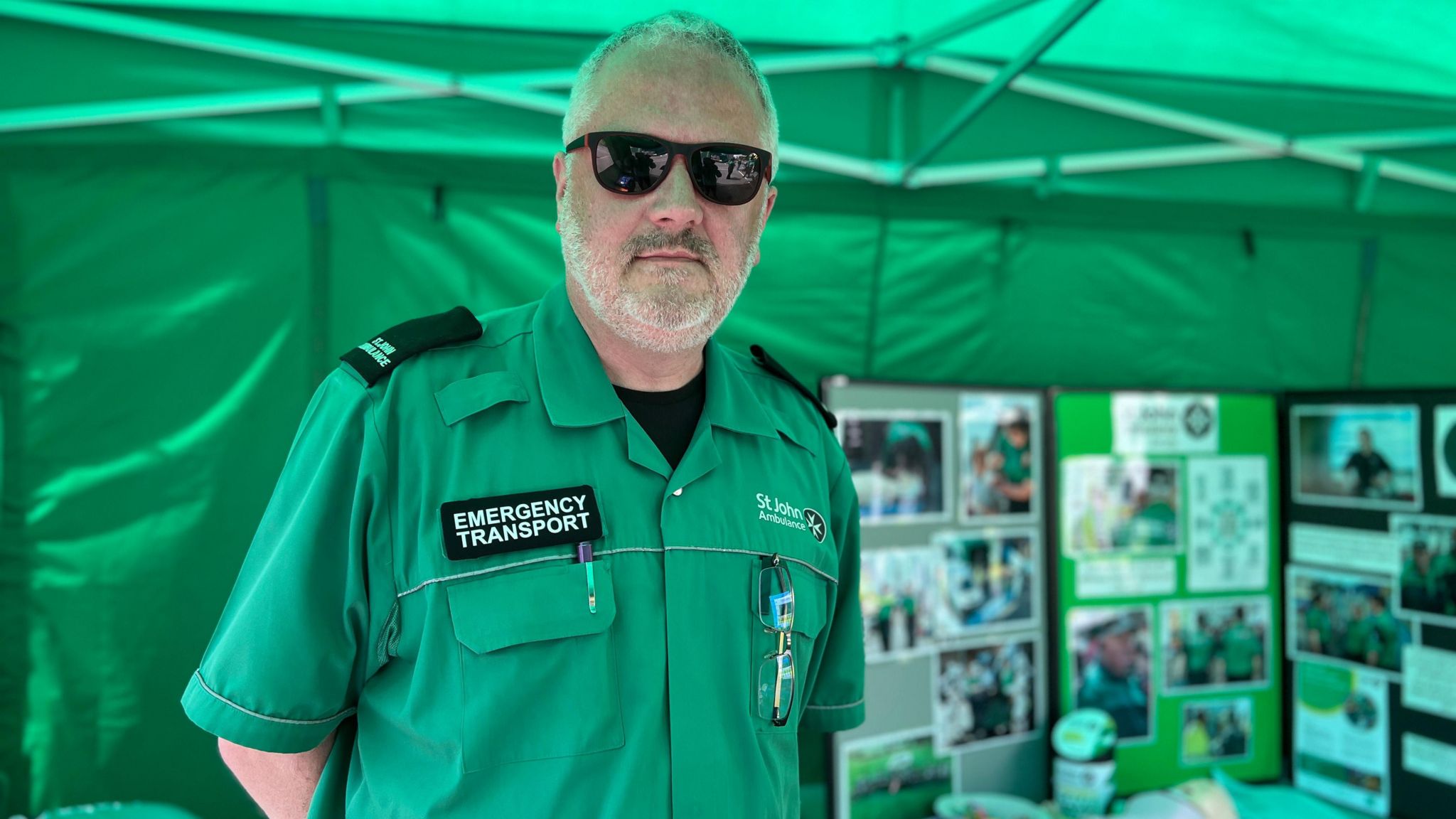 Mark looks at the camera while in his uniform and wearing sun glasses. He is in the charity's first aid tent with pictures behind him showing what the charity does