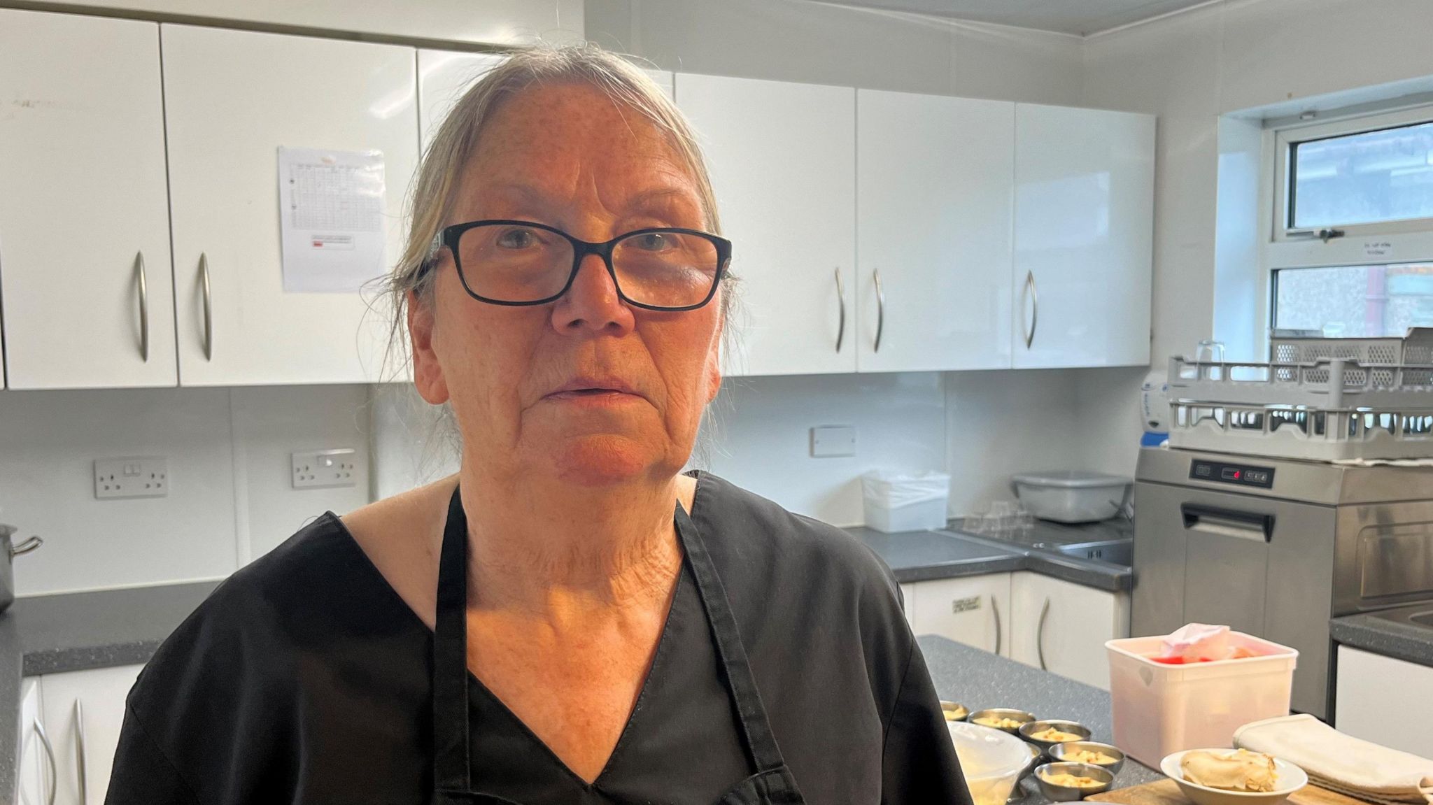 Cook Jan Rider, wearing glasses in care home kitchen with work surfaces and prepared food in the background