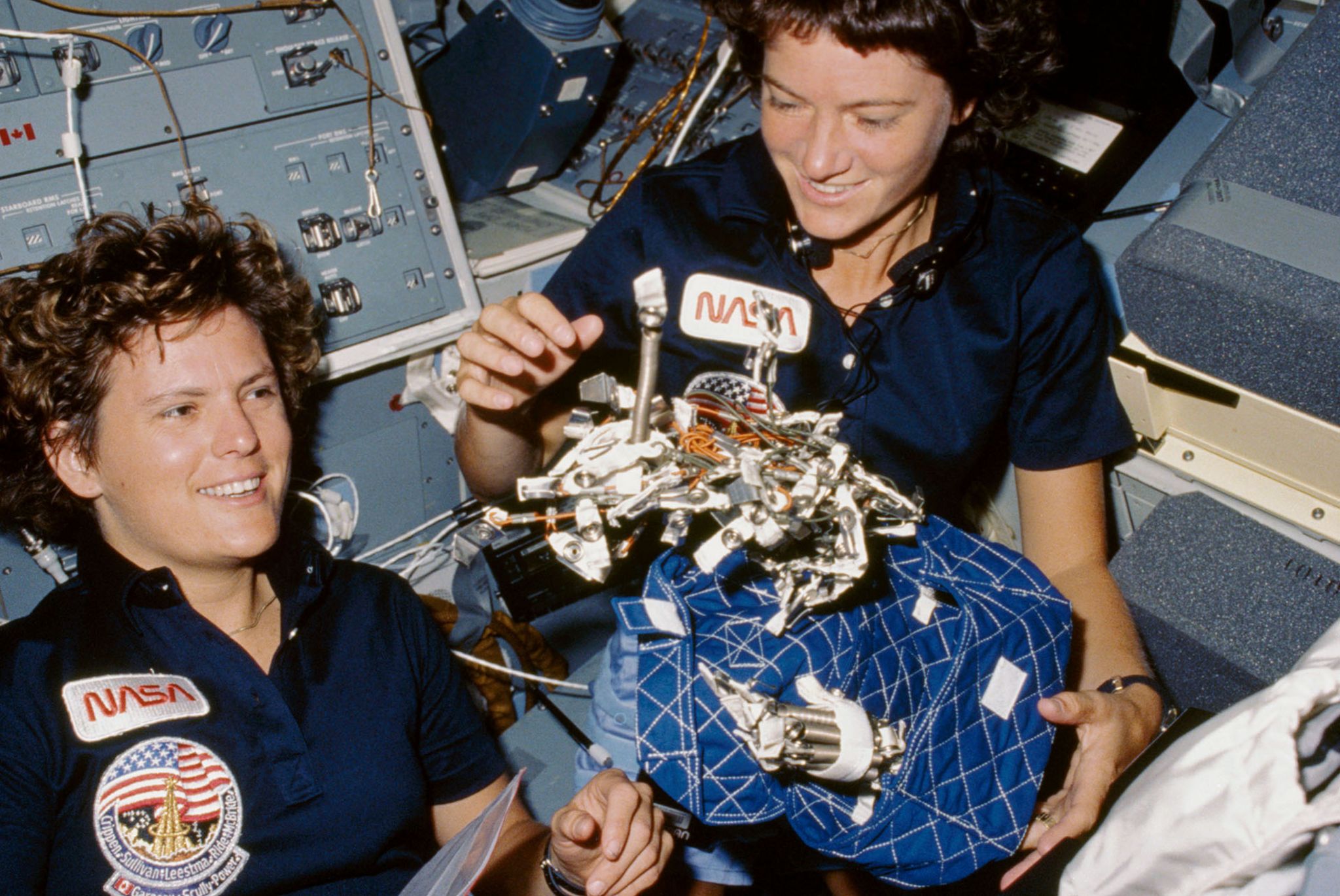 Astronauts Kathryn D. Sullivan, left, and Sally K. Ride display a "bag of worms." The "bag" is a sleep restraint and the majority of the "worms" are springs and clips used with the sleep restraint in its normal application