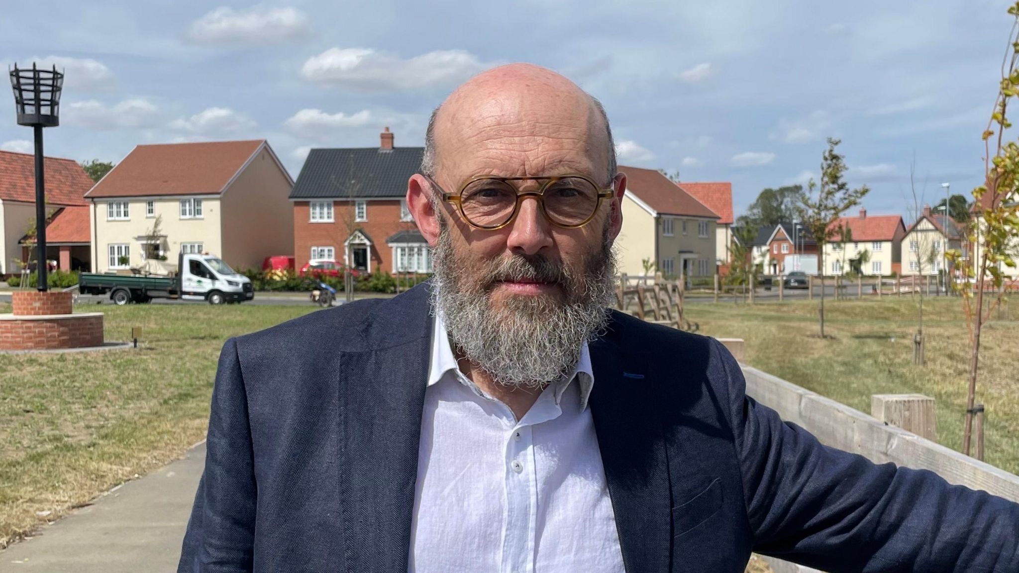 Councillor Andrew Stringer, wearing a dark blue jacket and white shirt, standing in front of the Beacon Green estate in Bacton. He wears round, plastic-rimmed glasses and a medium length greying beard.