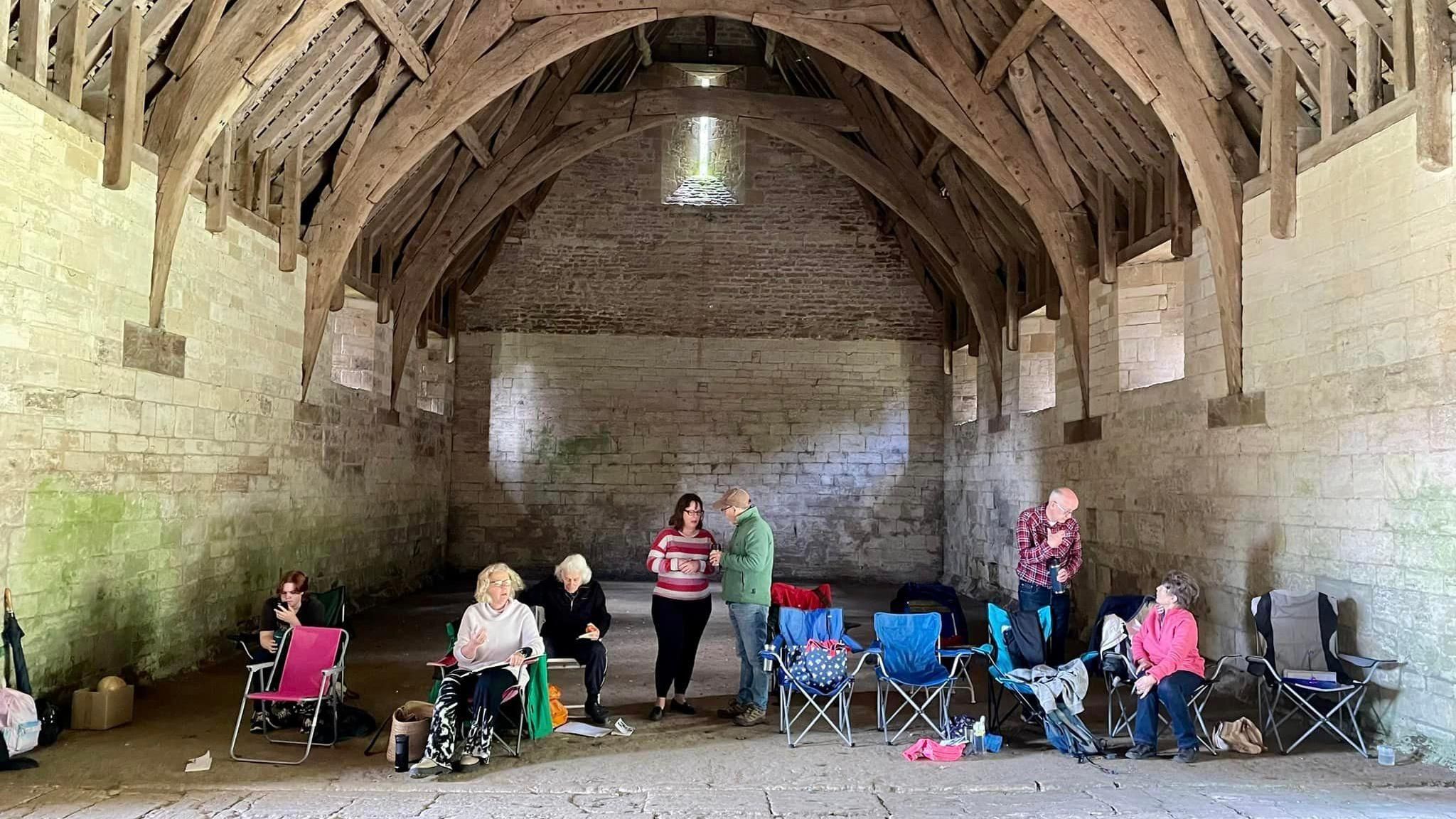 The Bradfordians Dramatic Society standing in the Tithe Barn. There are high arched wooden beam ceilings and stone slabs on the floor.