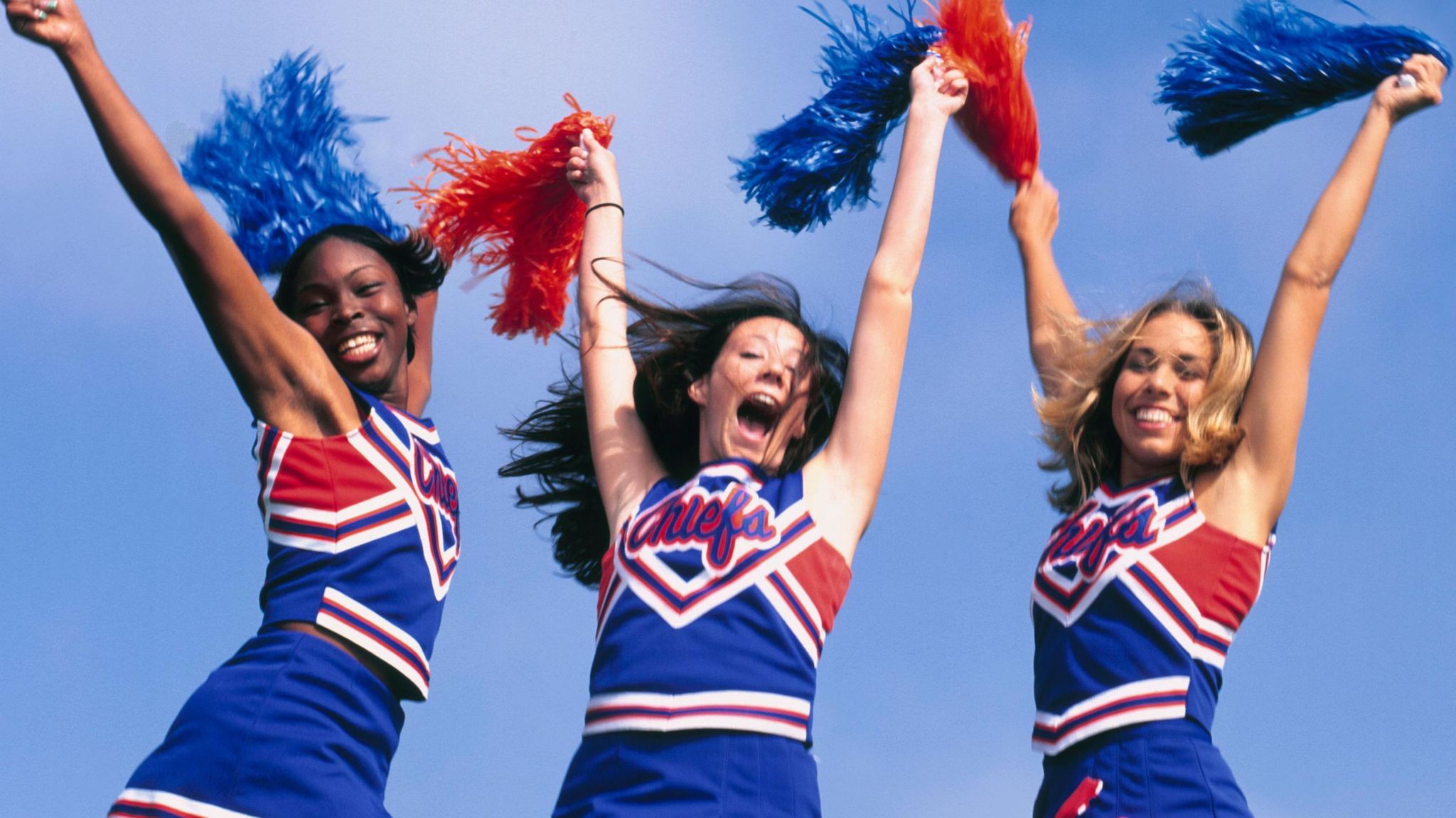 Three cheerleaders wearing blue uniforms jump while holding pom-poms