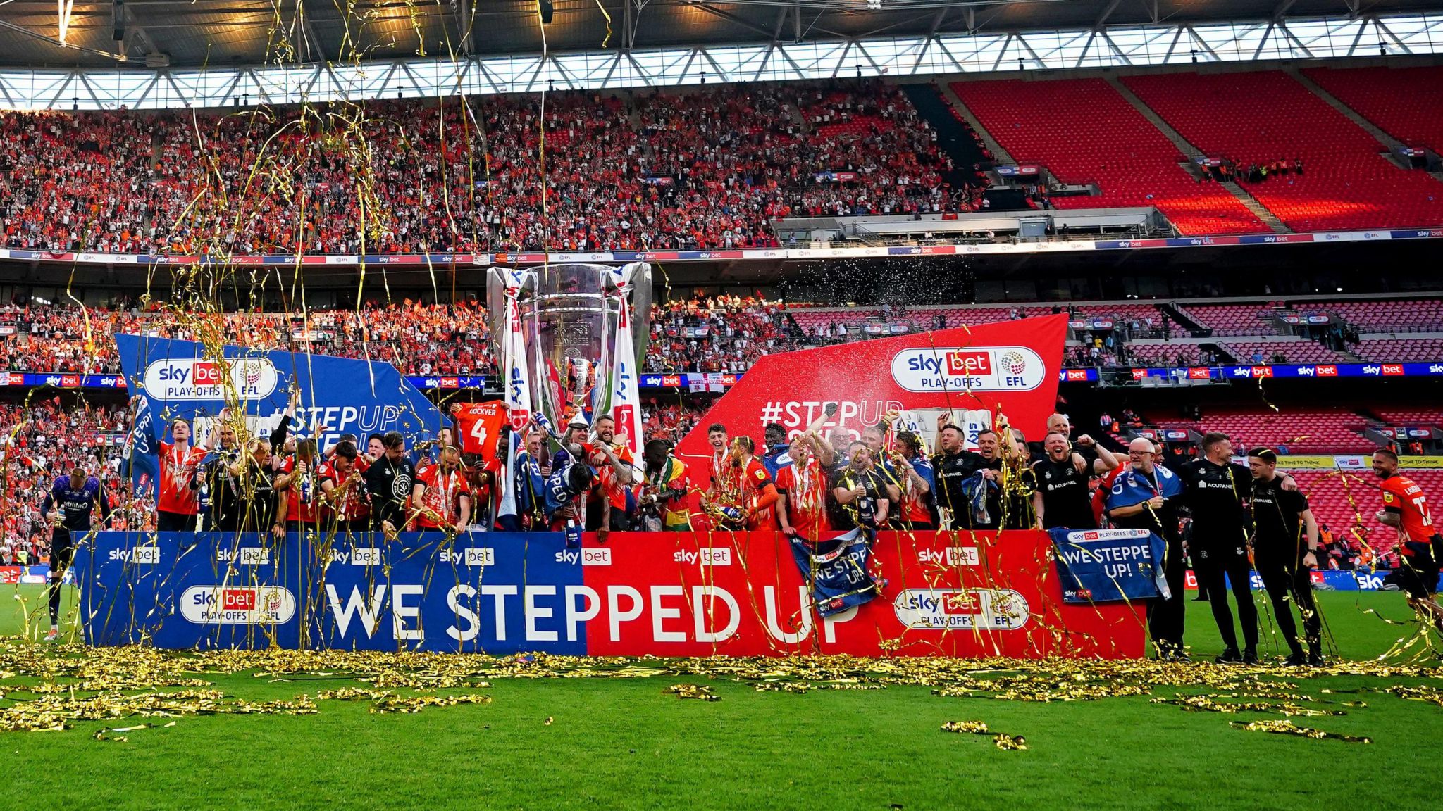 Luton's players and support staff are gathered in front of a board that has we stepped up in capped up white letters and Championship branding