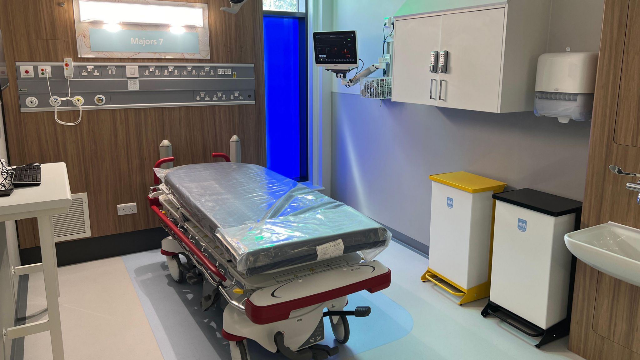 A treatment cubicle with a bed in the centre, a shelf and bins one one side, a laptop stand on the other and plug points in the background