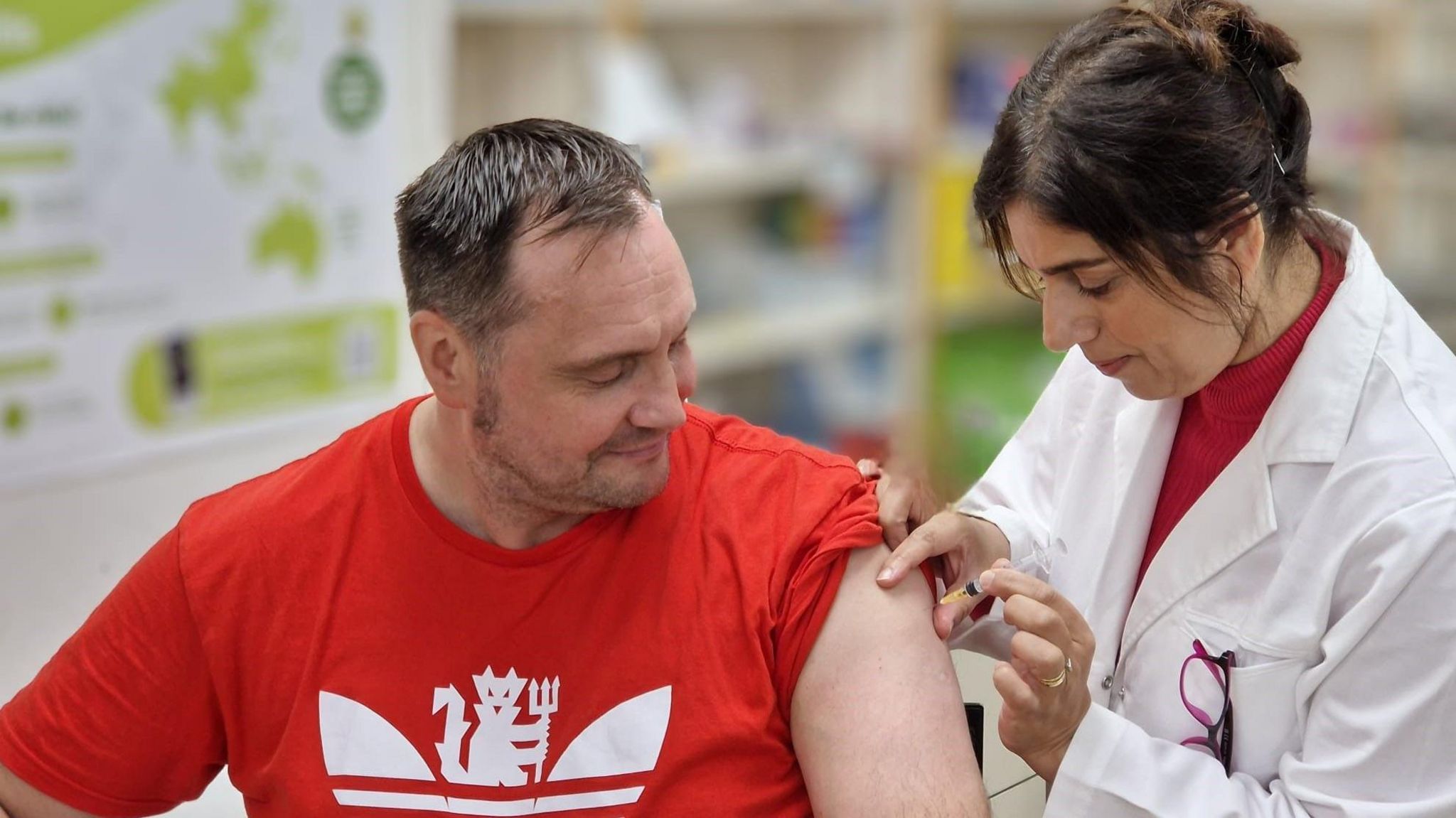 A woman in a white coat vaccinates a man sitting down and wearing a red t-shirt. 