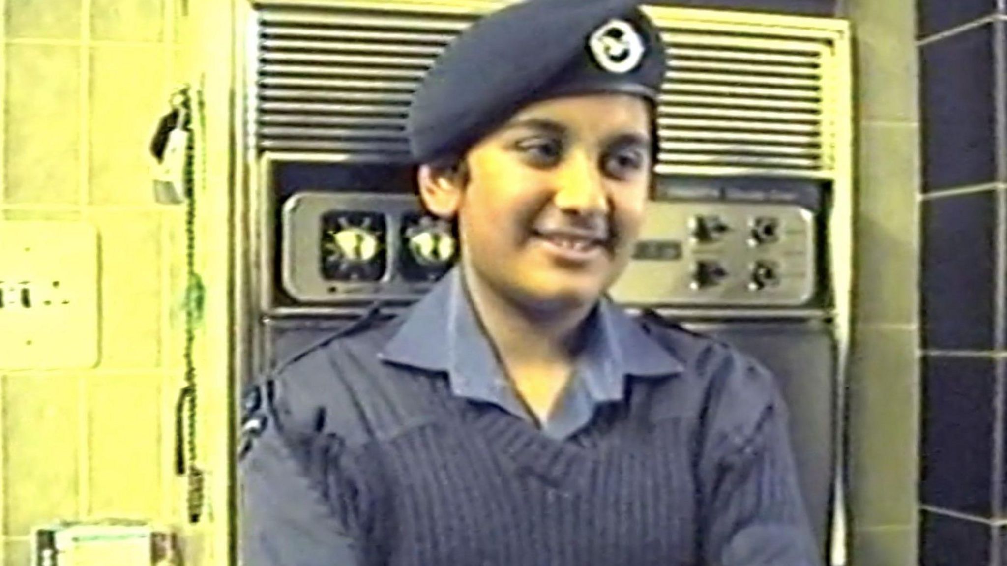 A young Ferhan in wearing a blue RAF cadet uniform and beret standing in front of a kitchen oven and smiling