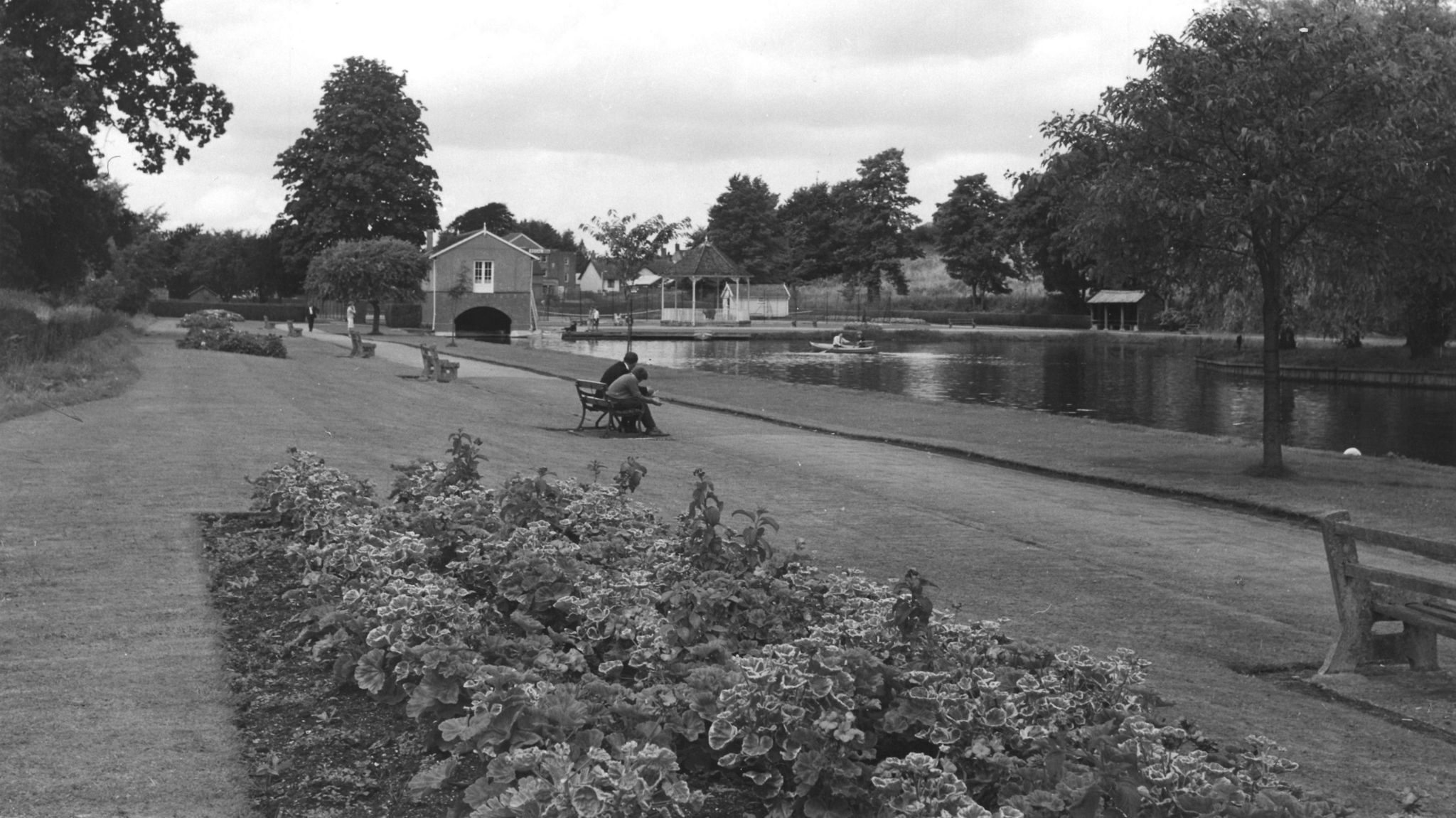 Black and white view of the park with lake, boathouse, flowerbeds and people sitting on benches