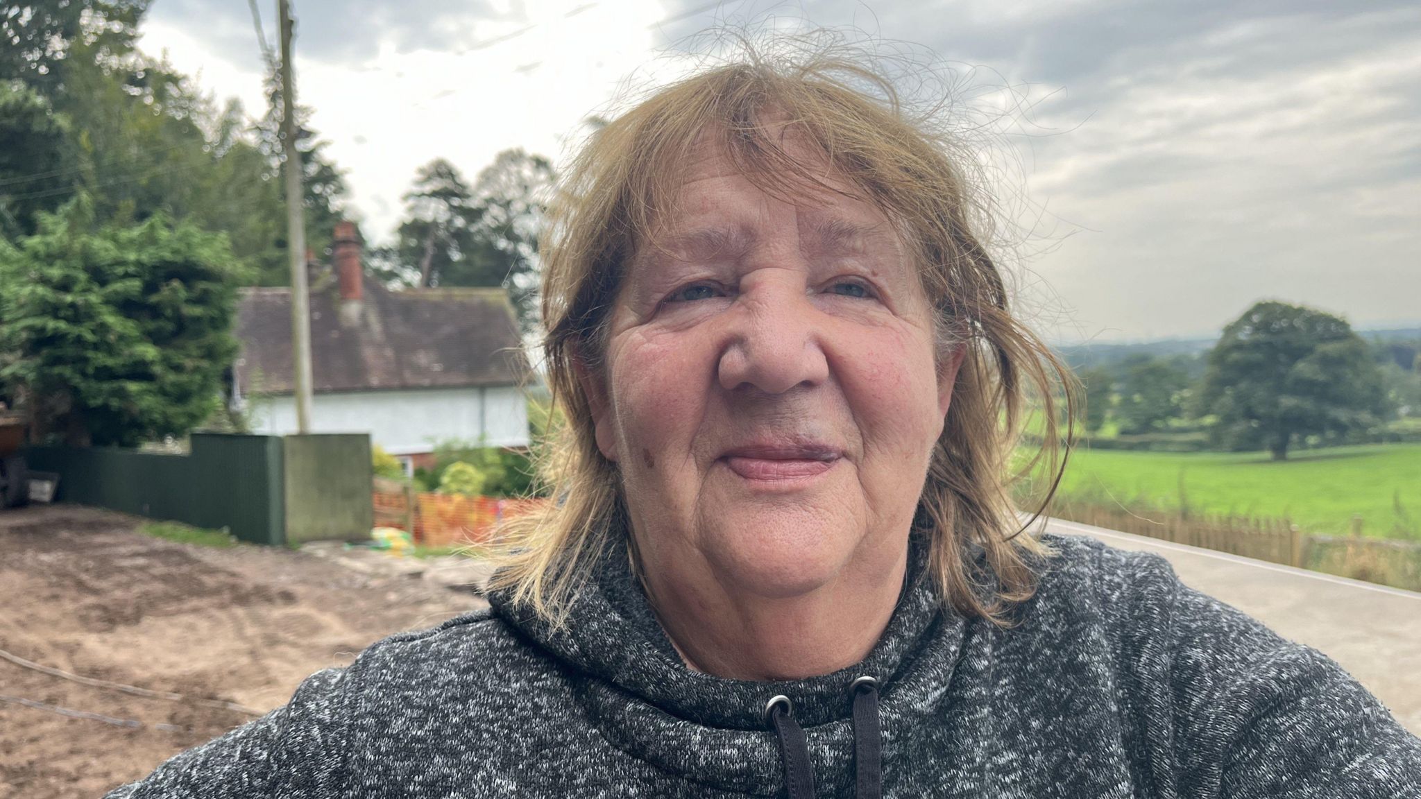 A woman with short brown hair smiles in to the camera. She is wearing a grey hoodie.