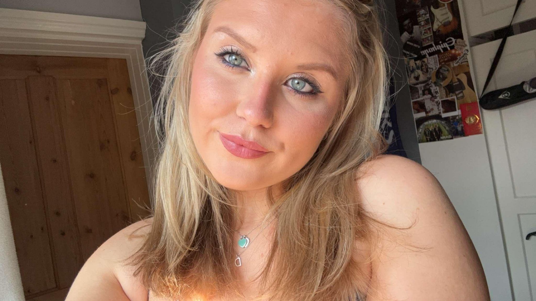 A selfie of a blonde girl with greenish, blue eyes in a bedroom