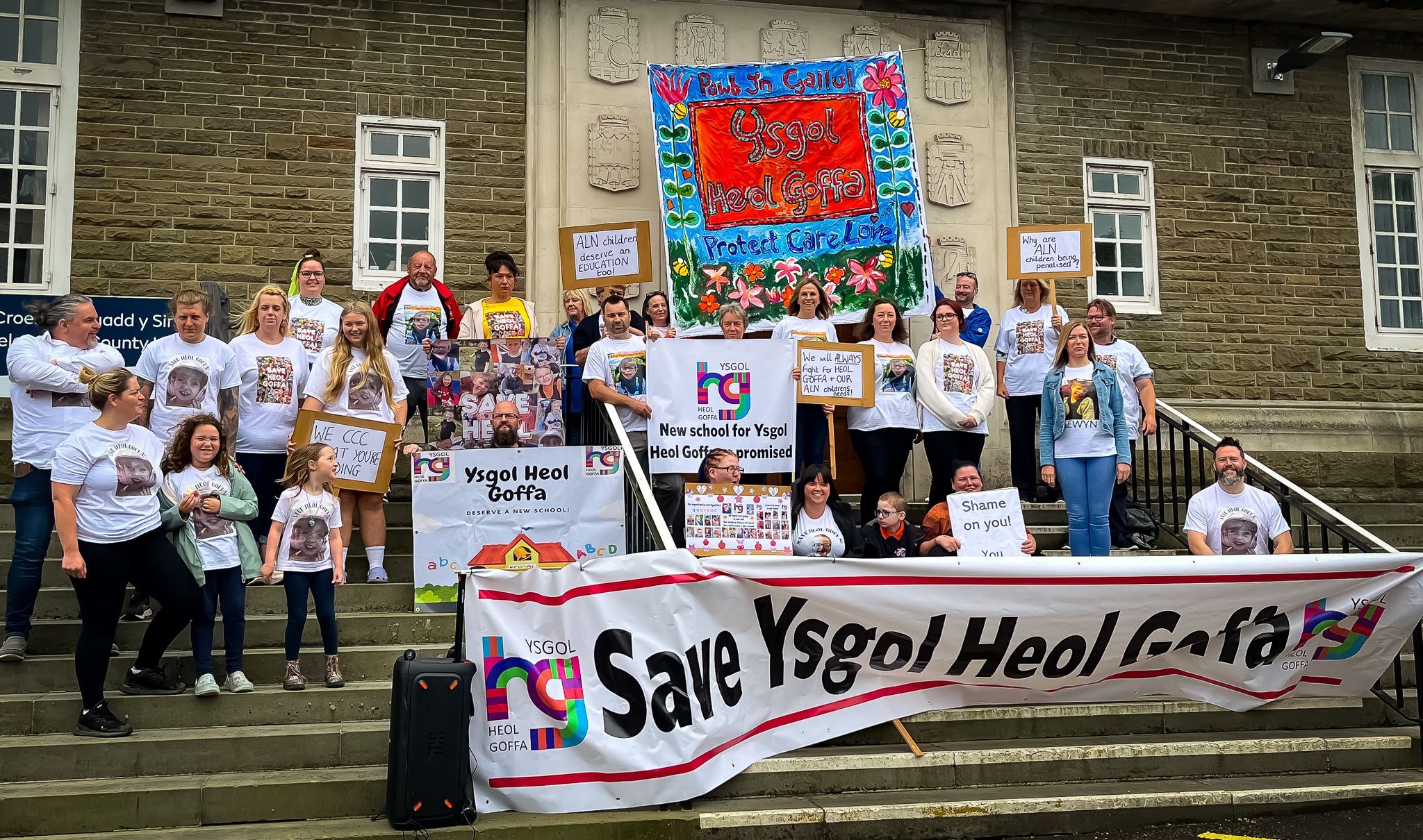 Protestsor wearing white shirts and holding a banner saying save ysgol heol goffa