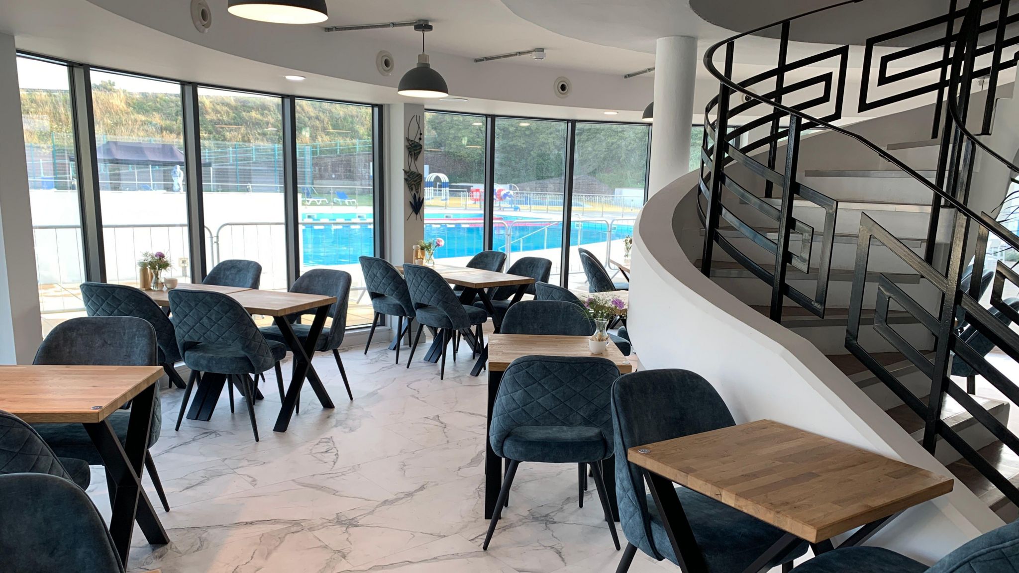 A dining room. There are dark wood tables with blue soft chair. A swimming pool is visible through large windows. There is a staircase to the right with black railings