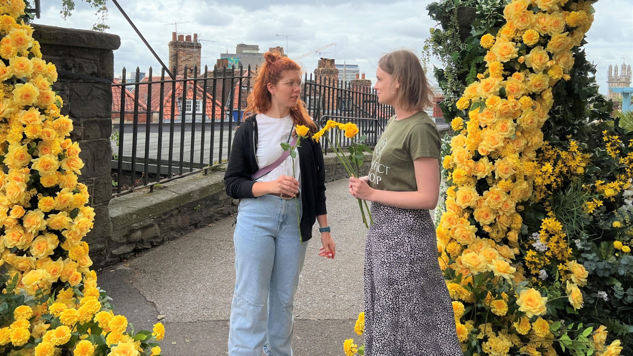 A woman holding a bunch of yellow flowers talking to another woman, who is holding one. They are surrounded either side by the flower structure