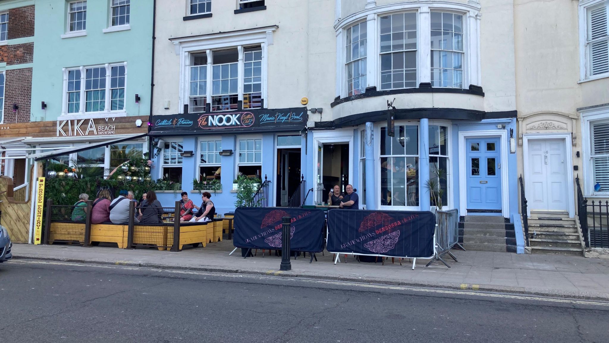 The Nook cocktail bar in Weymouth which has a black sign and door with steps leading up to the door. There is an outside seating are with wooden seats and tables at the front with a metal structure above it that looks like it could hold a canopy