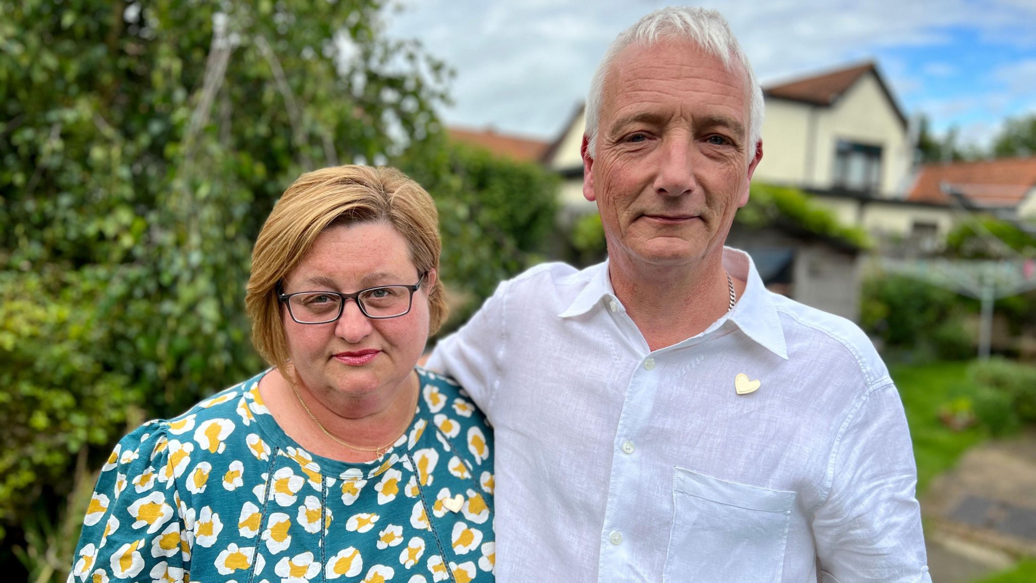 Lisa and James Woolnough standing in their garden looking into the camera. Lisa has a turquoise floral top and shoulder-length brunette hair and glasses. James has short, white hair and is wearing a white shirt.