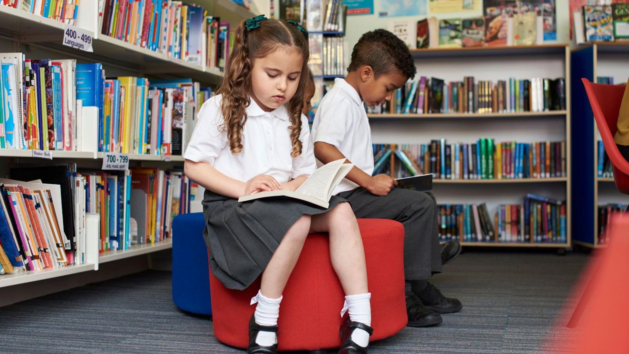 Two young children in school uniform reading in a library surrounded by books