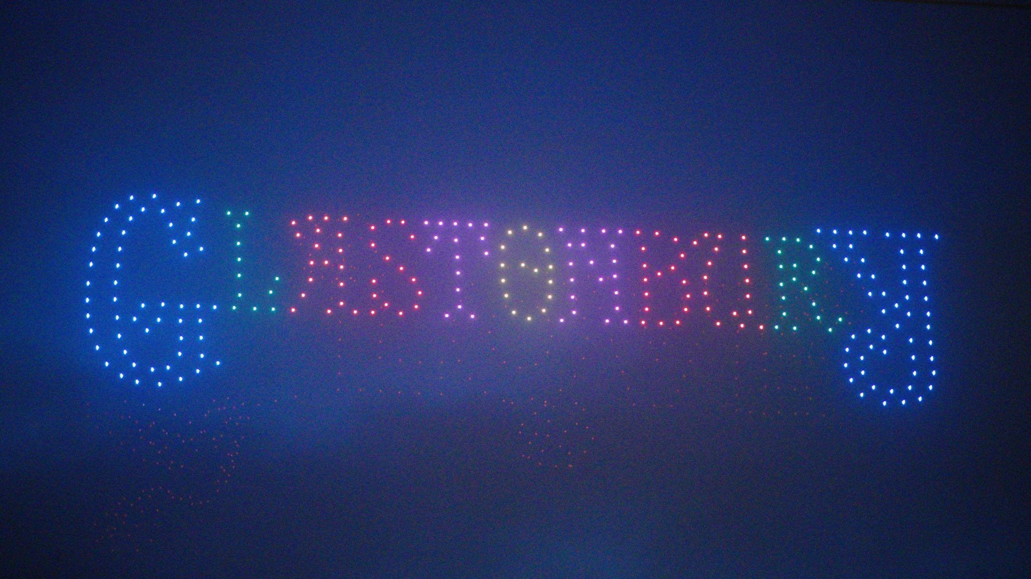 Image of a drone show at Glastonbury Festival. Glastonbury is spelled out with different coloured lights.