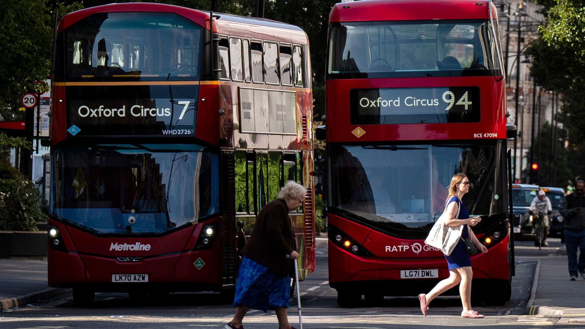 Two women, one with a walking stick, crossing the road while buses wait