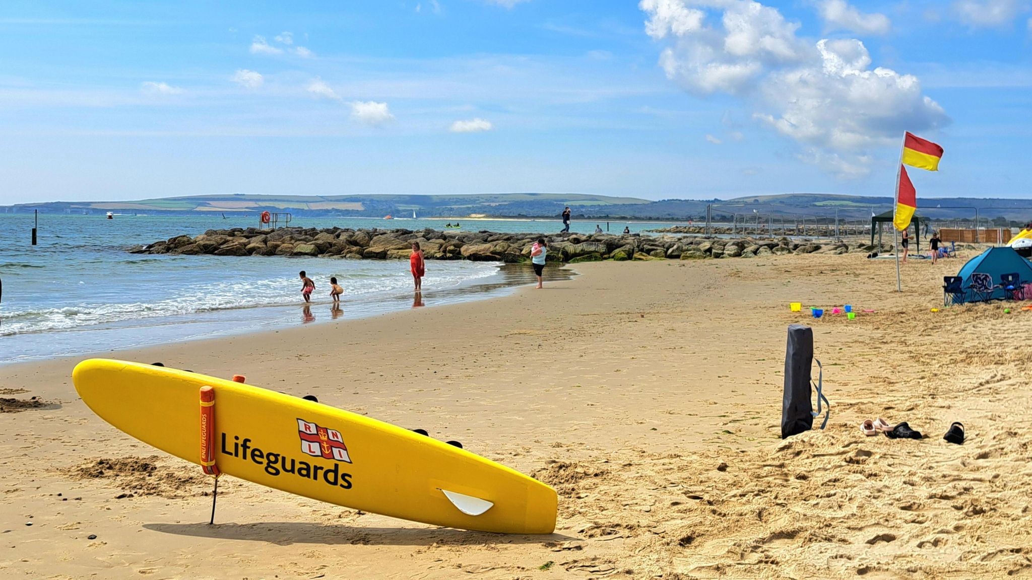 THURSDAY - A RNLI lifeboard is propped up ready for action if needed on beach with sweeping view of sand and sea, lifeguard's red and yellow flags fly to show safe swimming zone, with children playing at the water's edge being watched over by parents