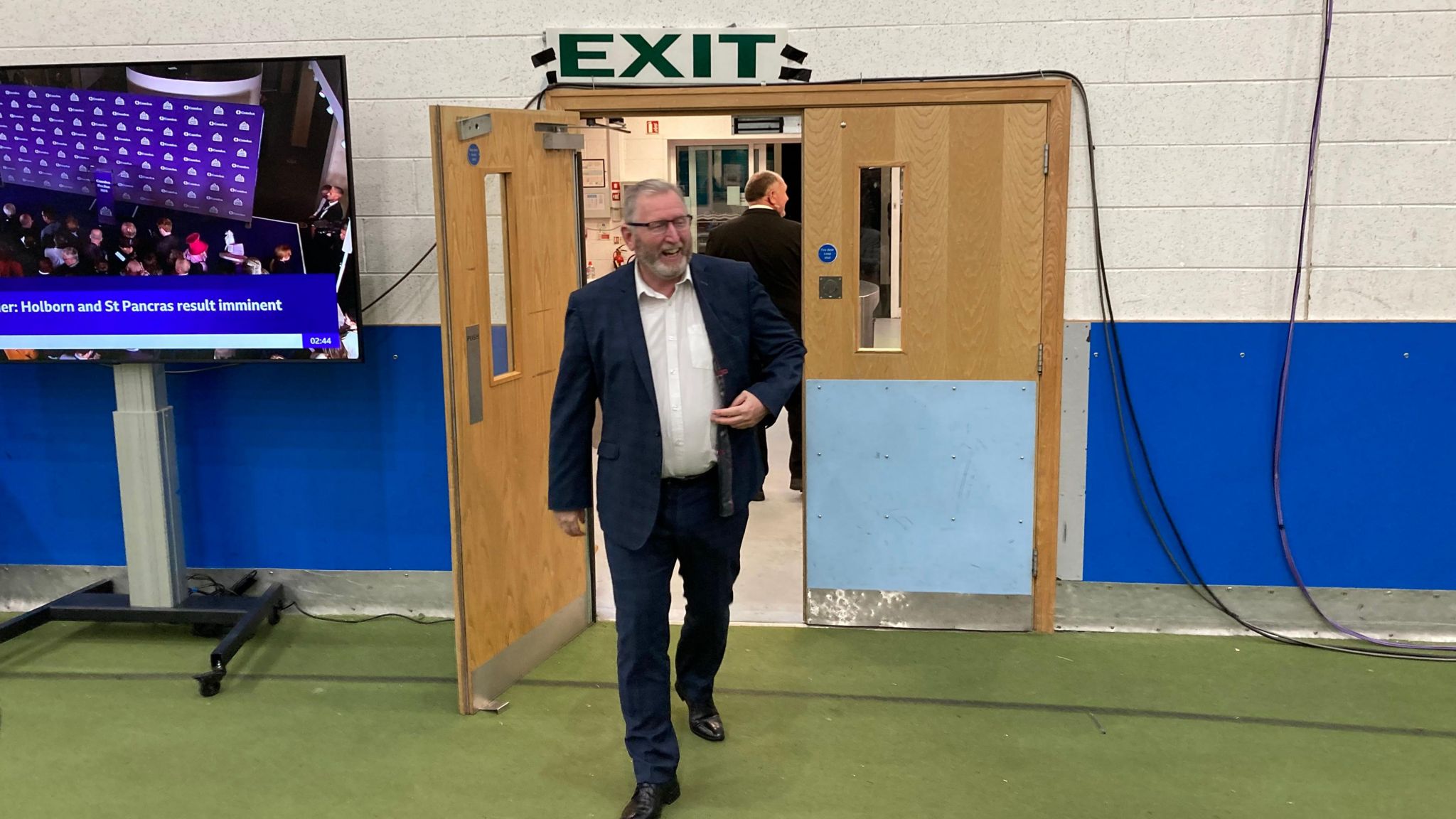 Doug Beattie in a dark suit walks through a door with an exit sign above it