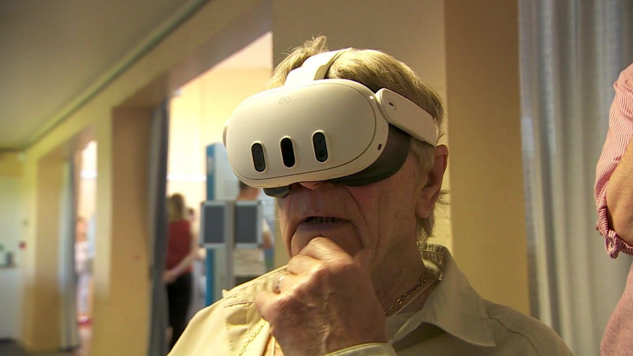 A man with grey hair is wearing a white virtual reality set. His hand is on his  chin, reflecting he ponders what he sees and his mouth is slightly ajar