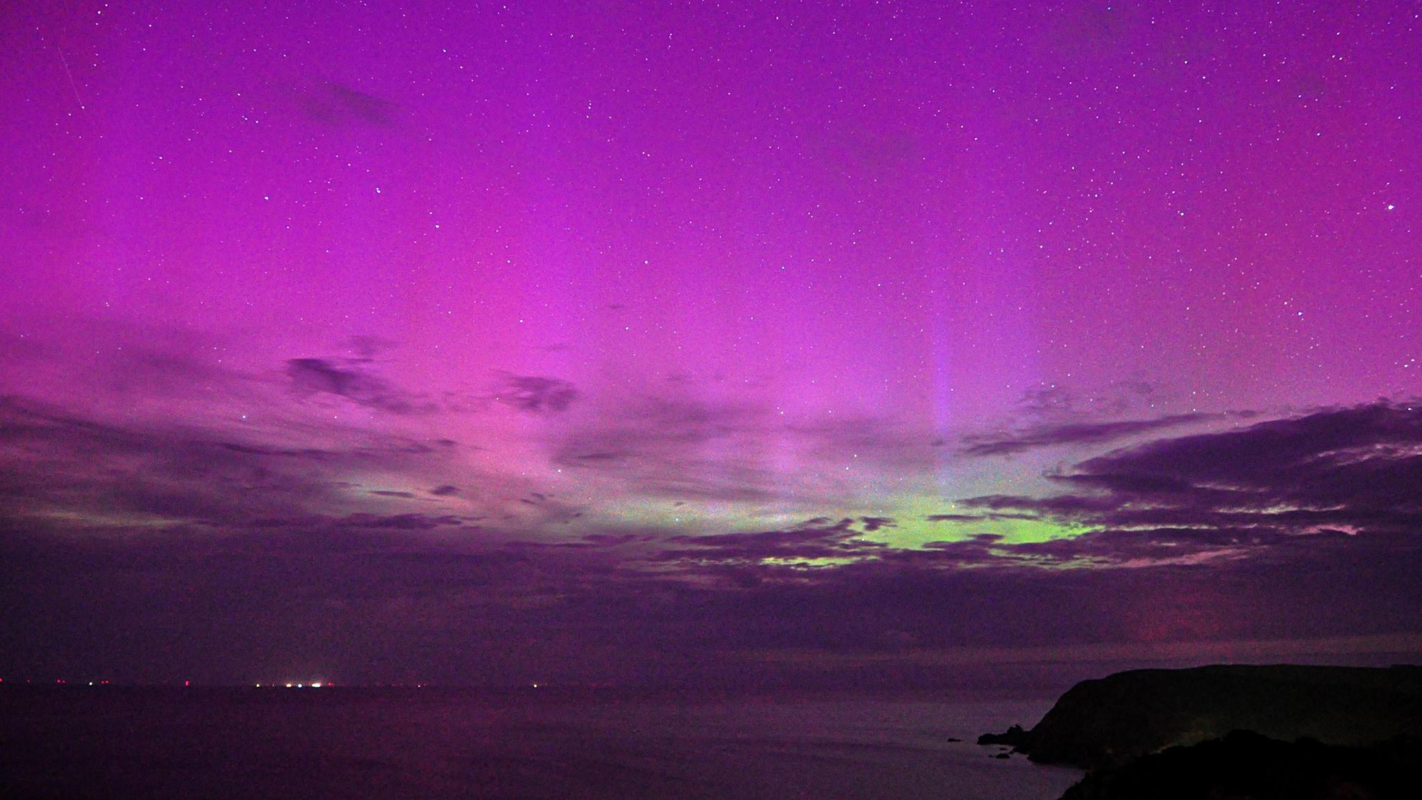 looking out to sea with some cloud in the distance and the northern lights shining bright in the sky.  Purples and greens peaking out of the cloud